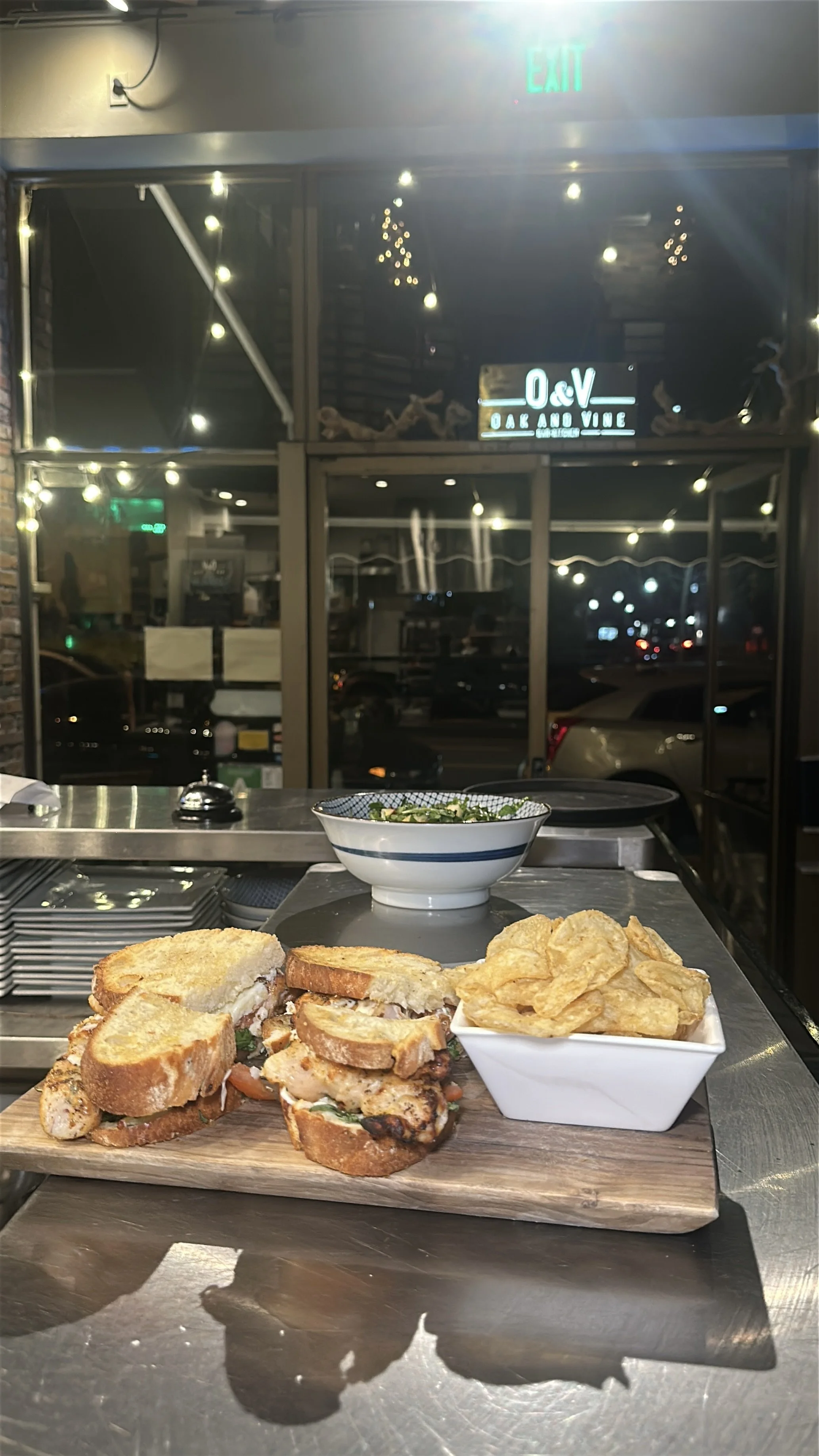 A wooden serving board with sandwiches, a bowl of potato chips, and a bowl of salad, set on a metal countertop inside a restaurant.