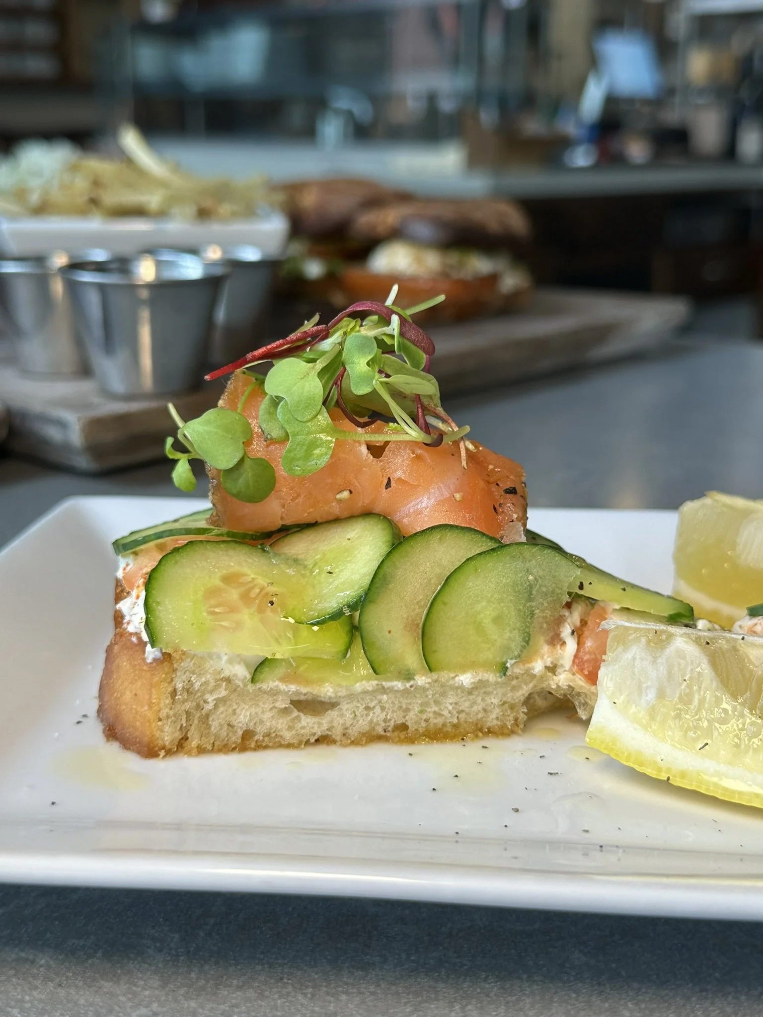 Close-up of a slice of bread topped with cucumber slices, smoked salmon, microgreens, and lemon wedges on a white plate.