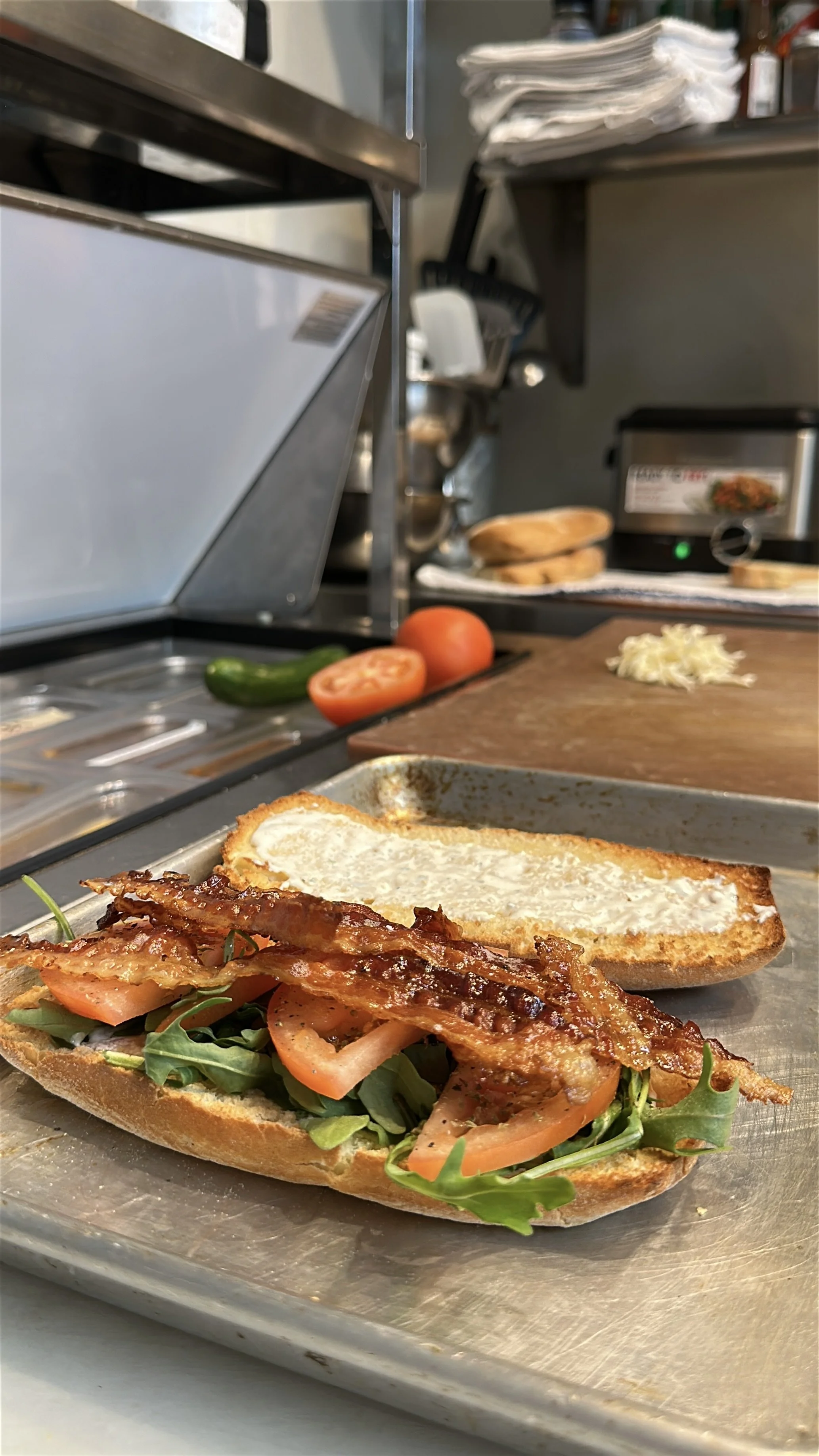 A half-assembled BLT sandwich on a stainless steel tray with fresh lettuce, tomato slices, crispy bacon, and toasted bread, with a smear of mayonnaise on the top slice.