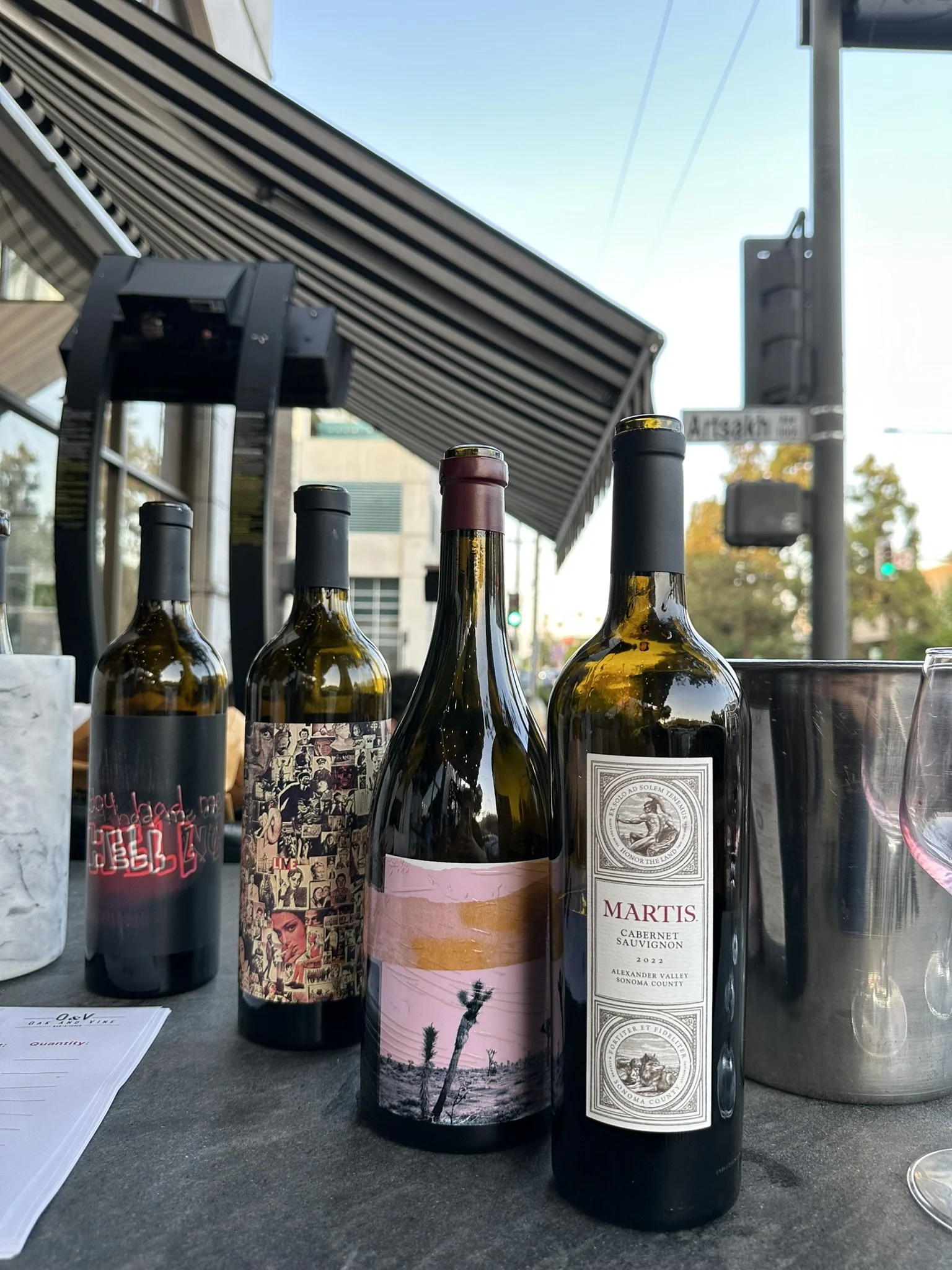 Four bottles of wine are lined up on a table outdoors, with a building, street signs, and trees in the background.