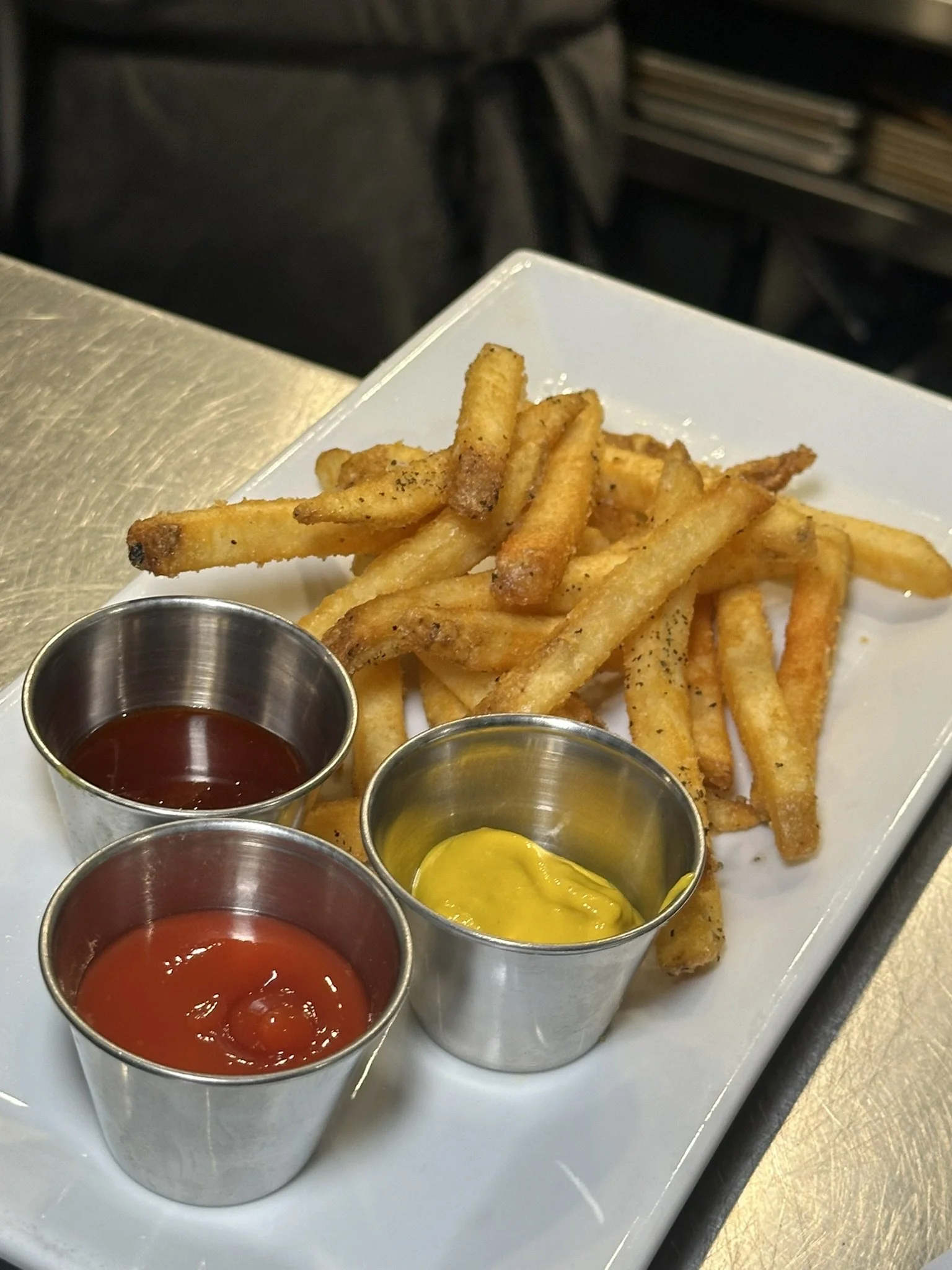 A plate of French fries served with ketchup and mustard in small metal cups.