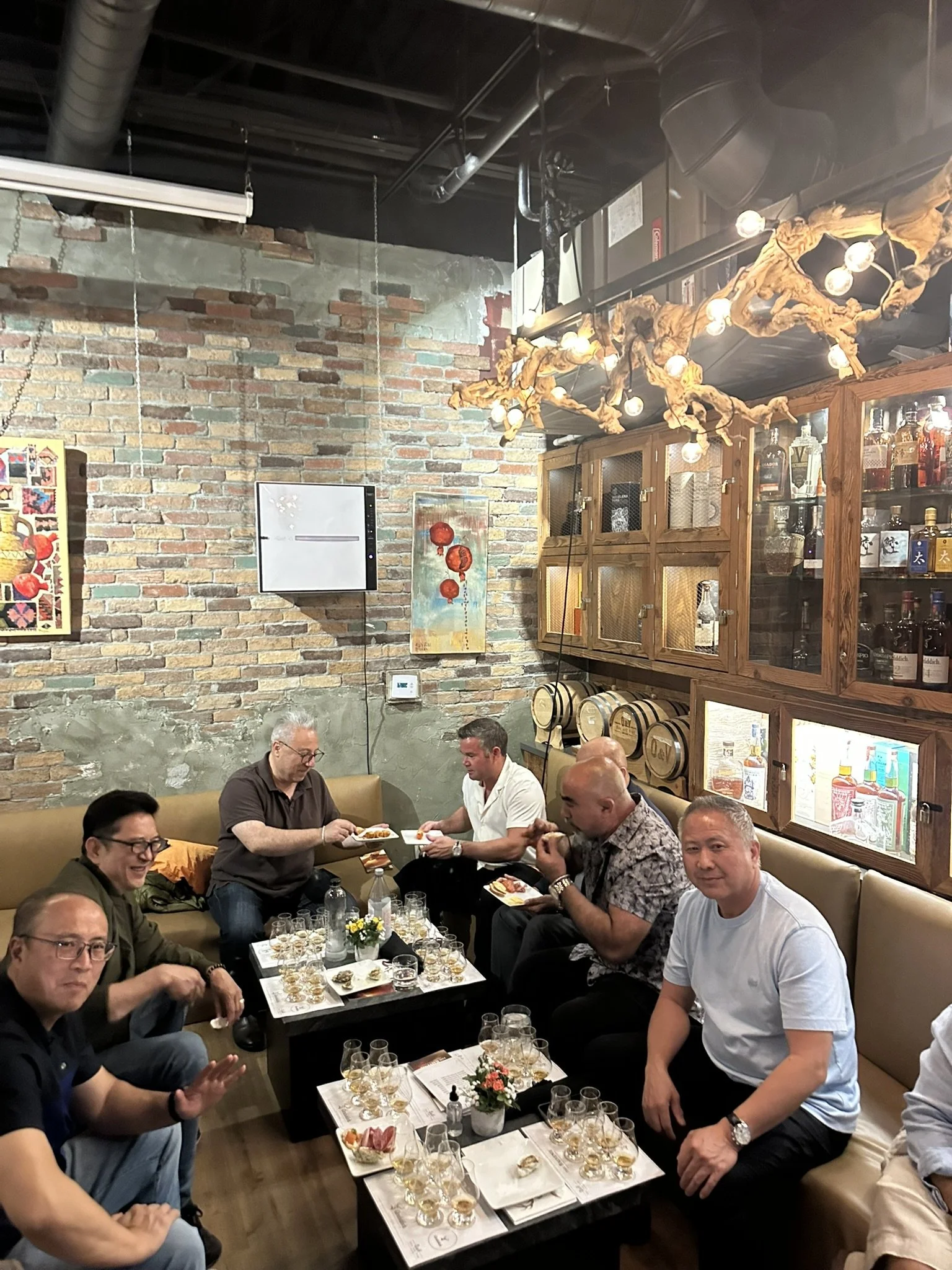 Group of people enjoying a social gathering in a cozy lounge or bar, seated around tables with glasses and snacks, with exposed brick walls and decorative lighting.