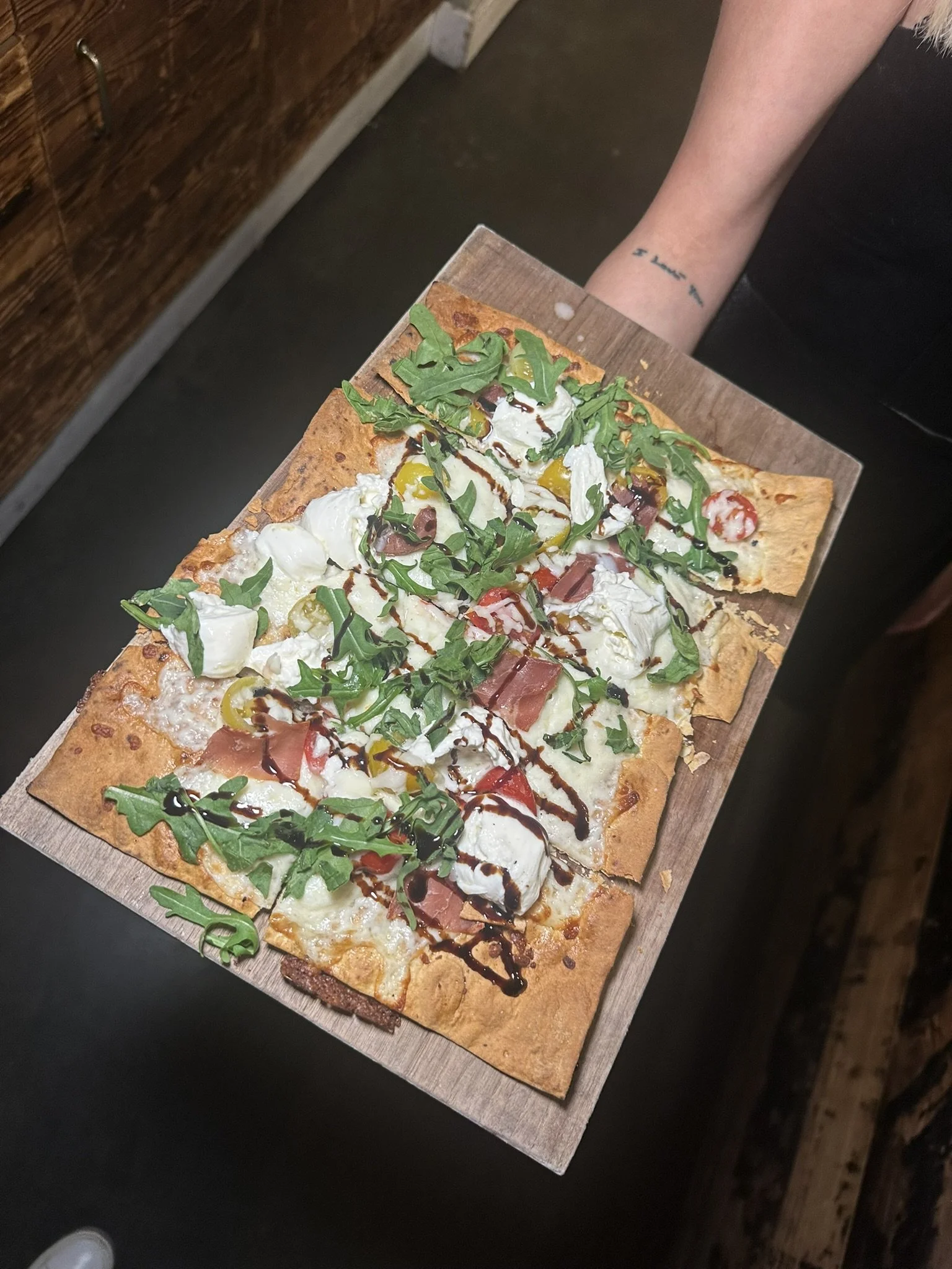 A person holding a rectangular wooden tray with a flatbread pizza topped with arugula, cherry tomatoes, mozzarella cheese, slices of prosciutto, yellow peppers, and drizzled with balsamic glaze and balsamic vinegar.
