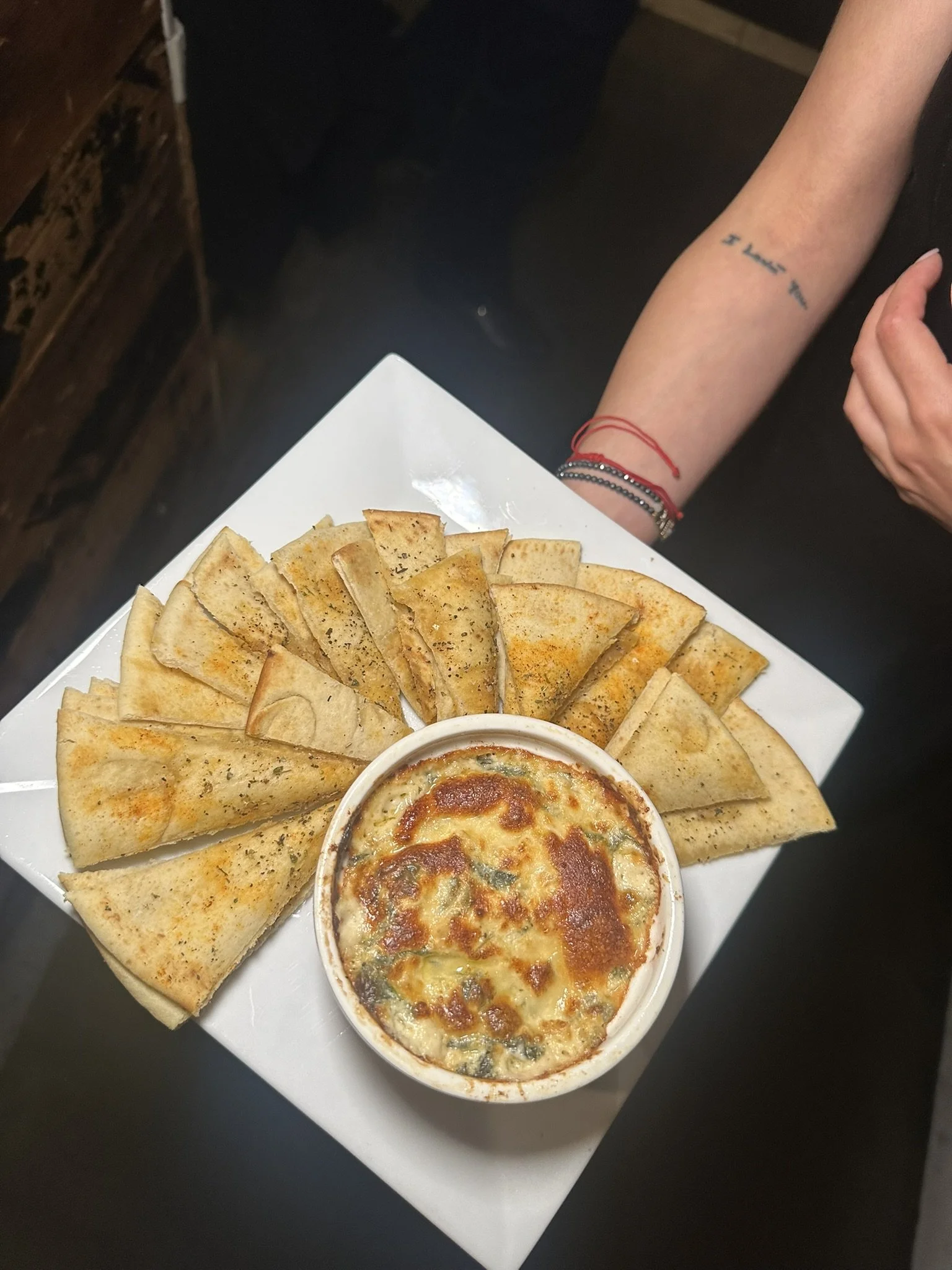 A white square plate holds sliced garlic bread arranged around a small bowl of baked spinach and cheese dip, with melted cheese on top.