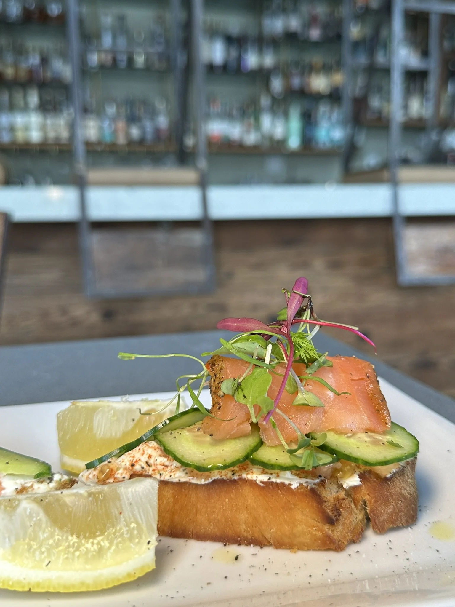 An open-faced smoked salmon sandwich with cucumber slices, microgreens, and lemon wedges on a white plate.