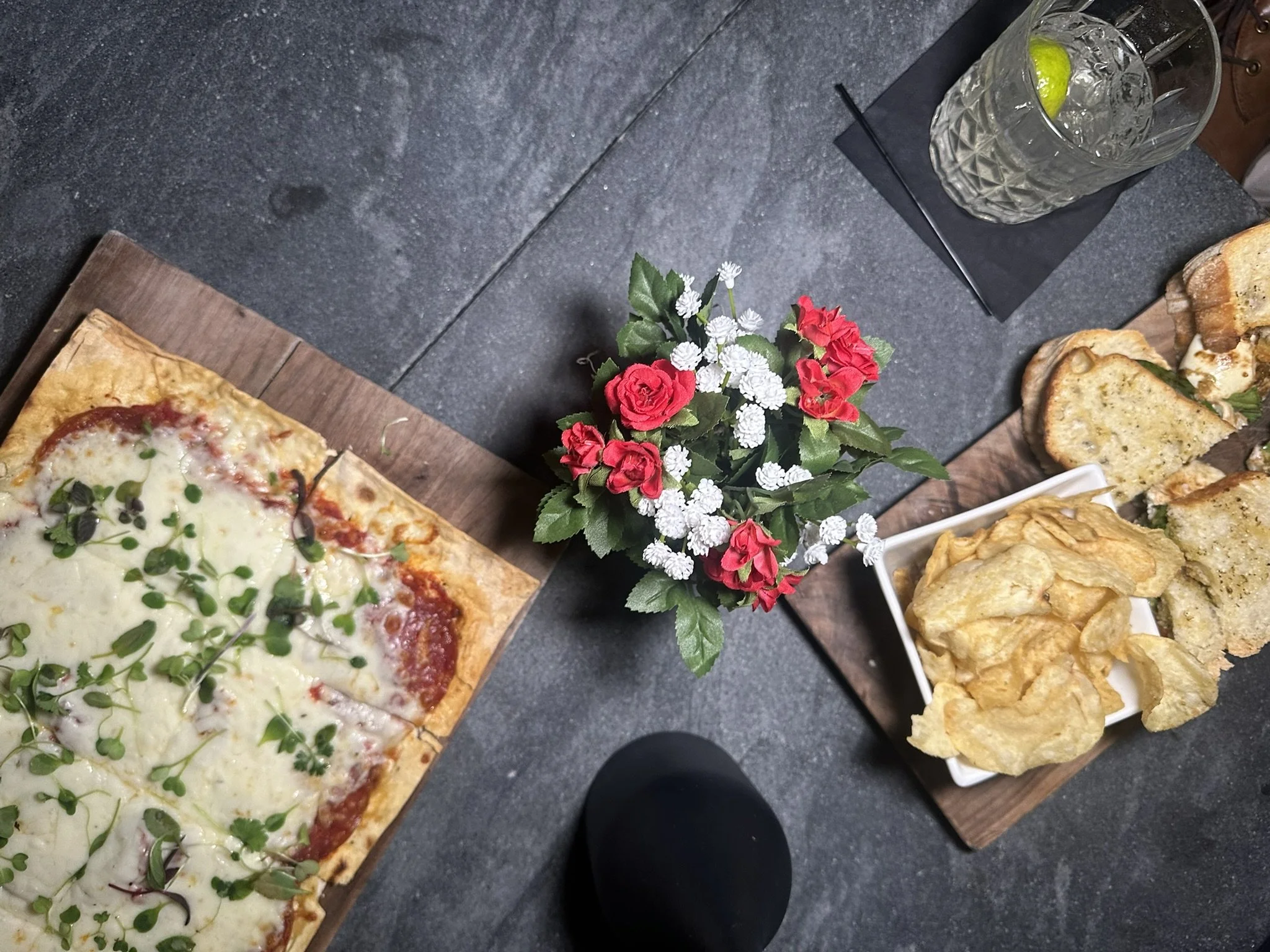Top-down view of a dining table with a plant in the center, a glass of water with a lime wedge, a pizza on a wooden board, and a small plate of potato chips and bread slices on the side.