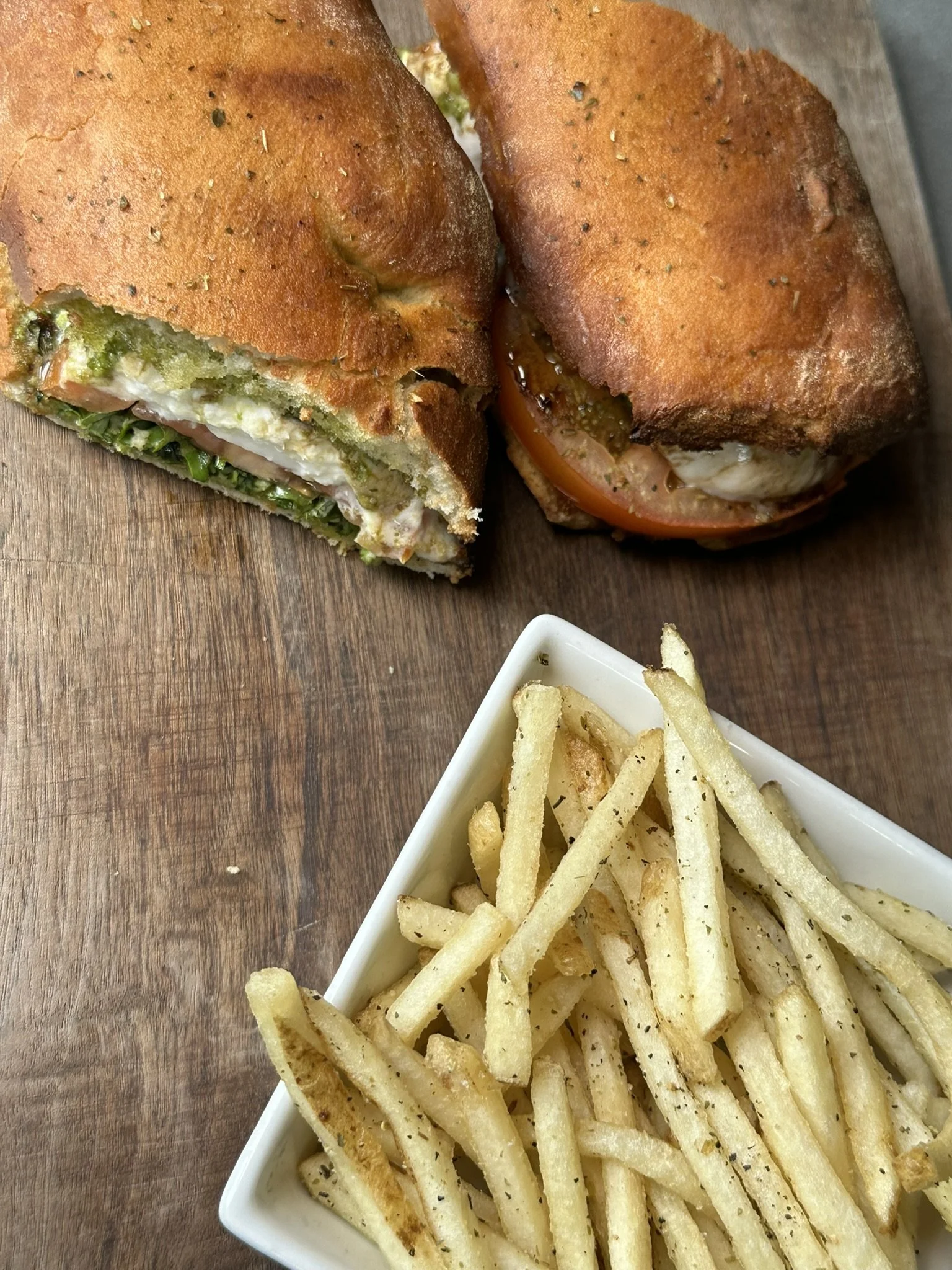 Half of a sandwich with green vegetables and cheese, a halved tomato with mozzarella, and a bowl of French fries on a wooden surface.