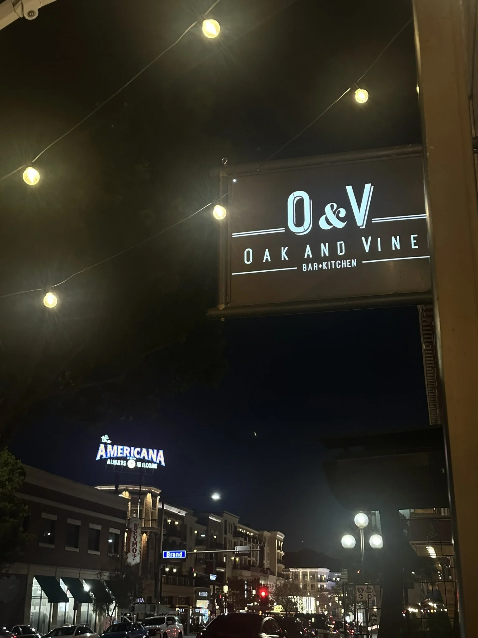 Night view of a busy city street with illuminated signs, including a prominent sign for 'O&V Oak and Vine Bar & Kitchen' and another for 'the Americana' with a welcoming message.