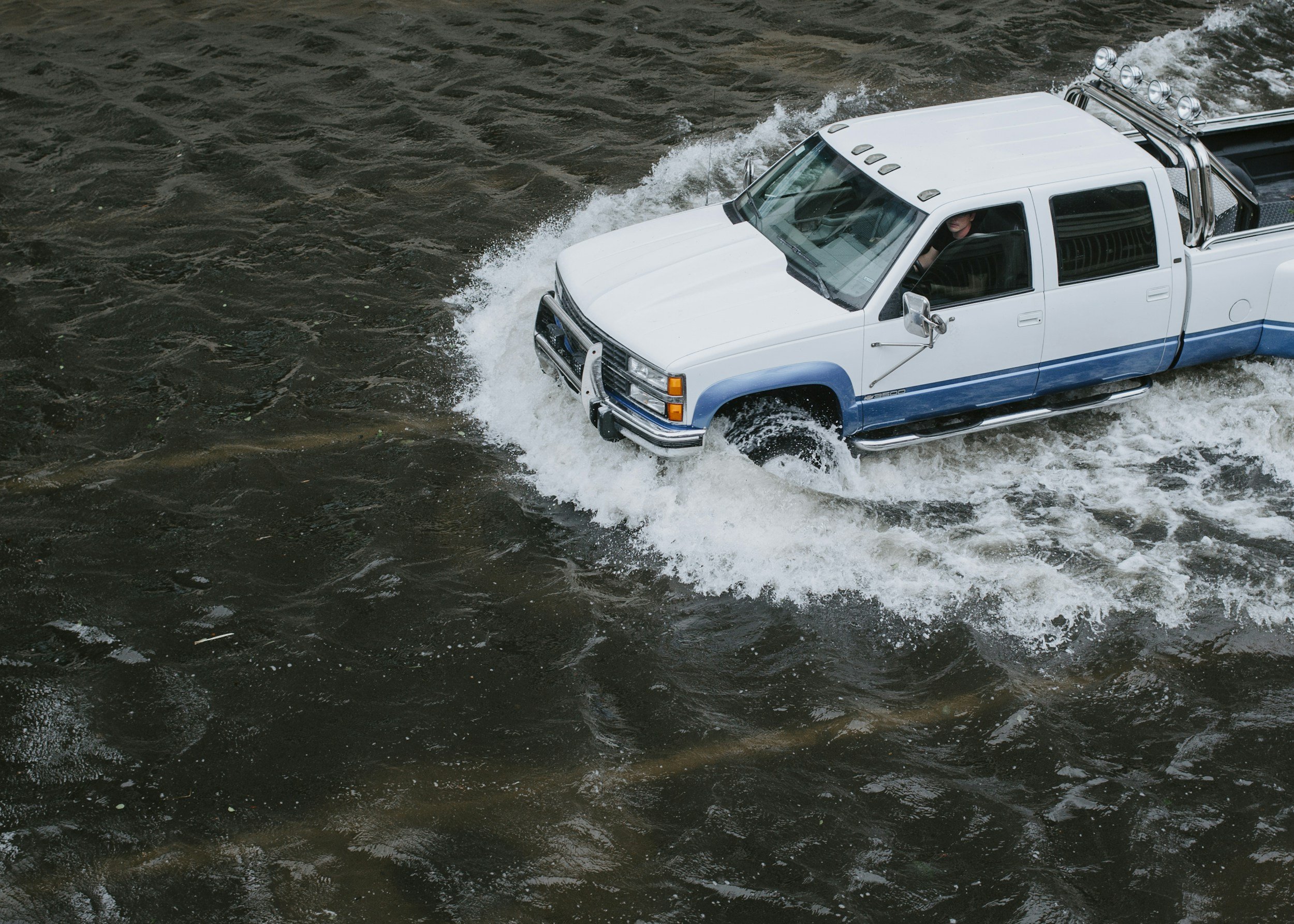 A white and blue pickup truck driving through water on a flooded road, creating splashes.
