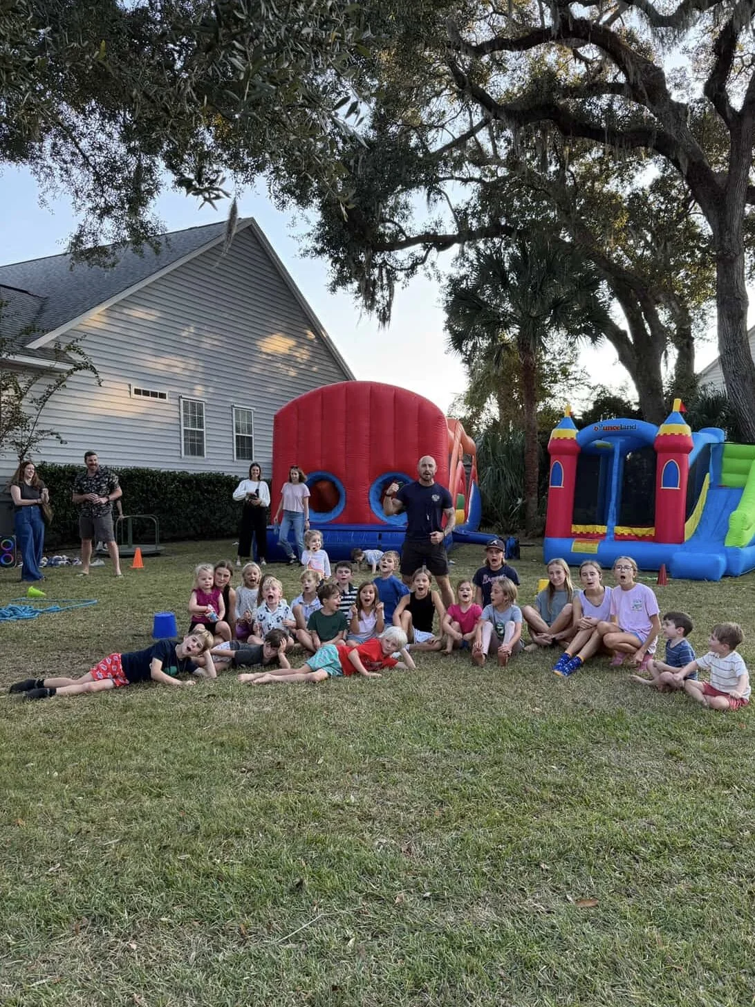 Group of children and adults at a backyard birthday party with inflatable bounce houses, on a grassy lawn in front of a house with trees.