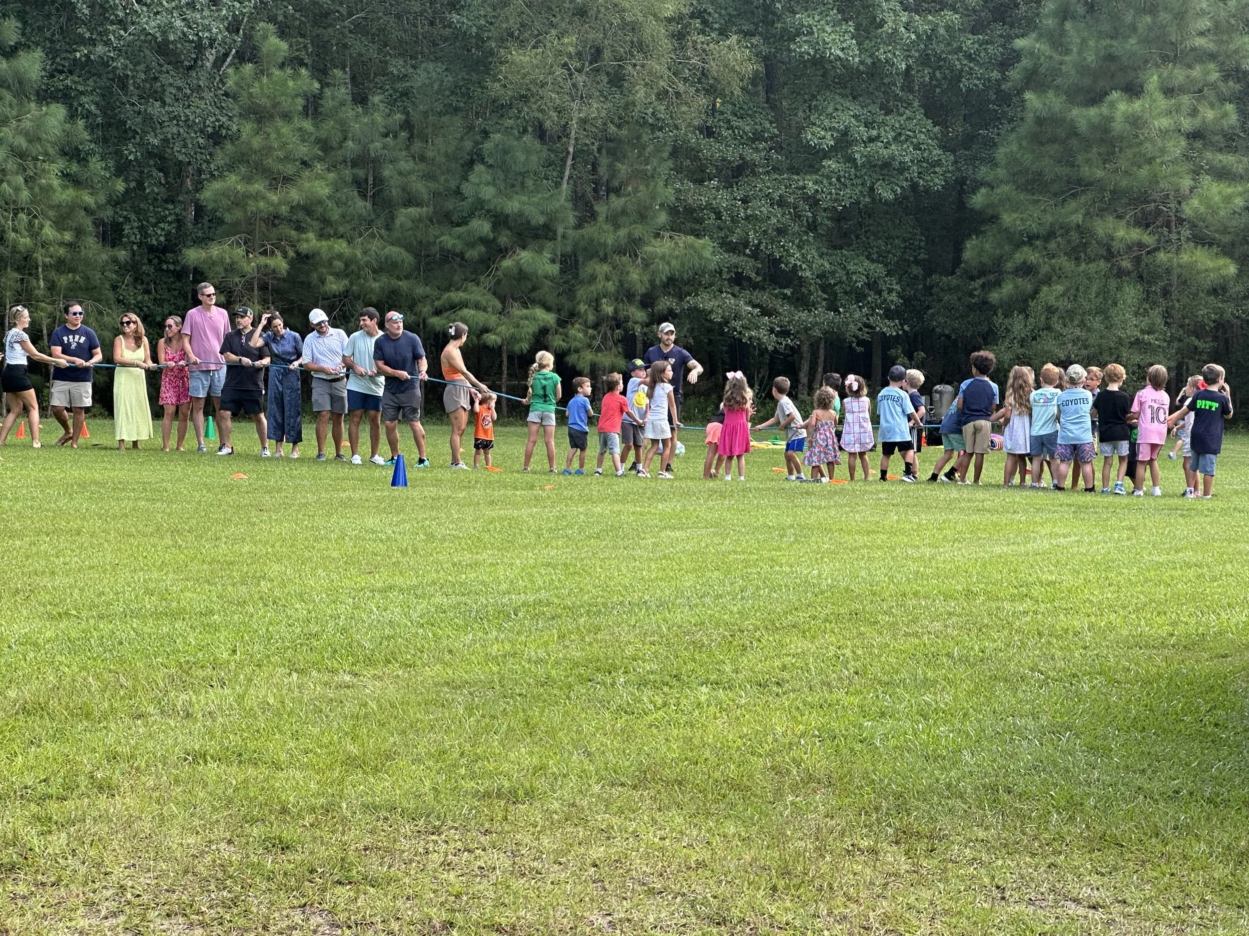 Group of children and adults participating in a tug-of-war game outdoors on a grassy field with trees in the background.