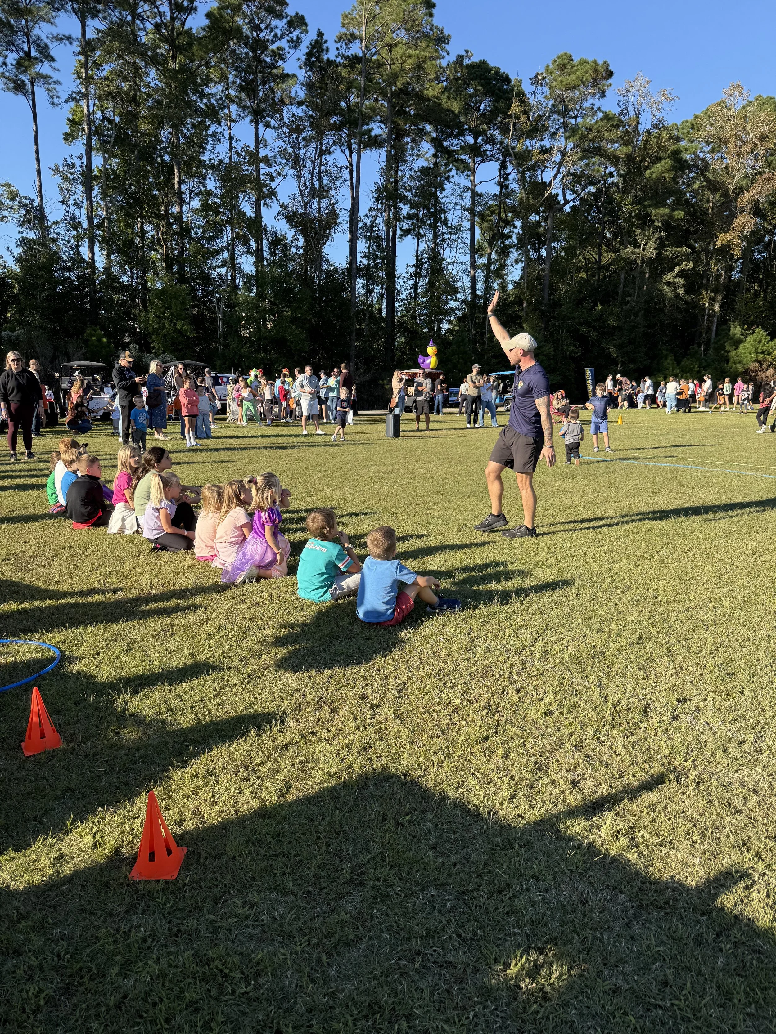 A group of children seated on the grass watching a man perform a trick on a field during a community event. There are many people in the background, some walking and some gathered around stands and tents. The setting is outdoors with clear blue skies and tall trees.