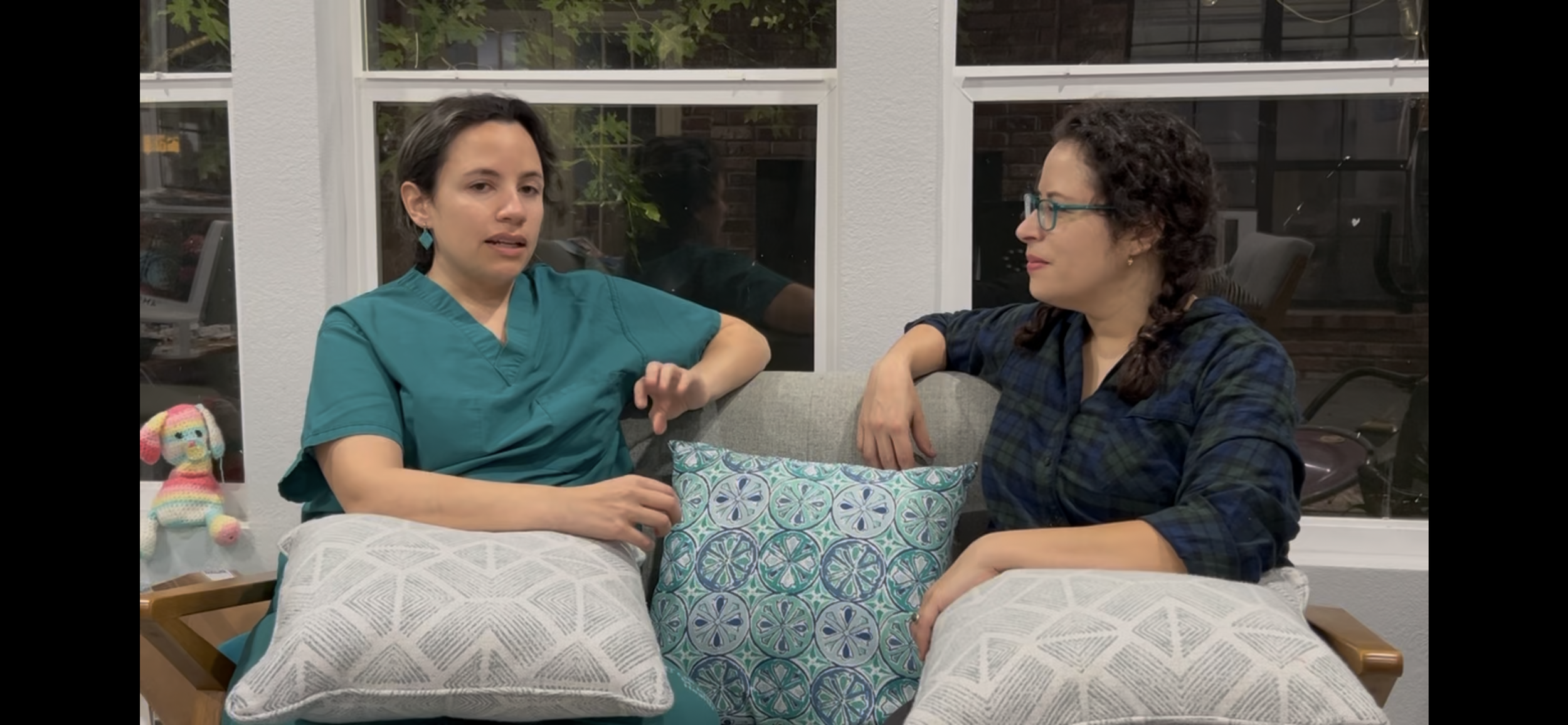 Romy and her sister, Dr. Adrianna Goldberg, sitting on a couch, engaged in conversation. Dr. Adriana Goldberg is wearing teal scrubs, and Natalia is wearing glasses and a plaid shirt.