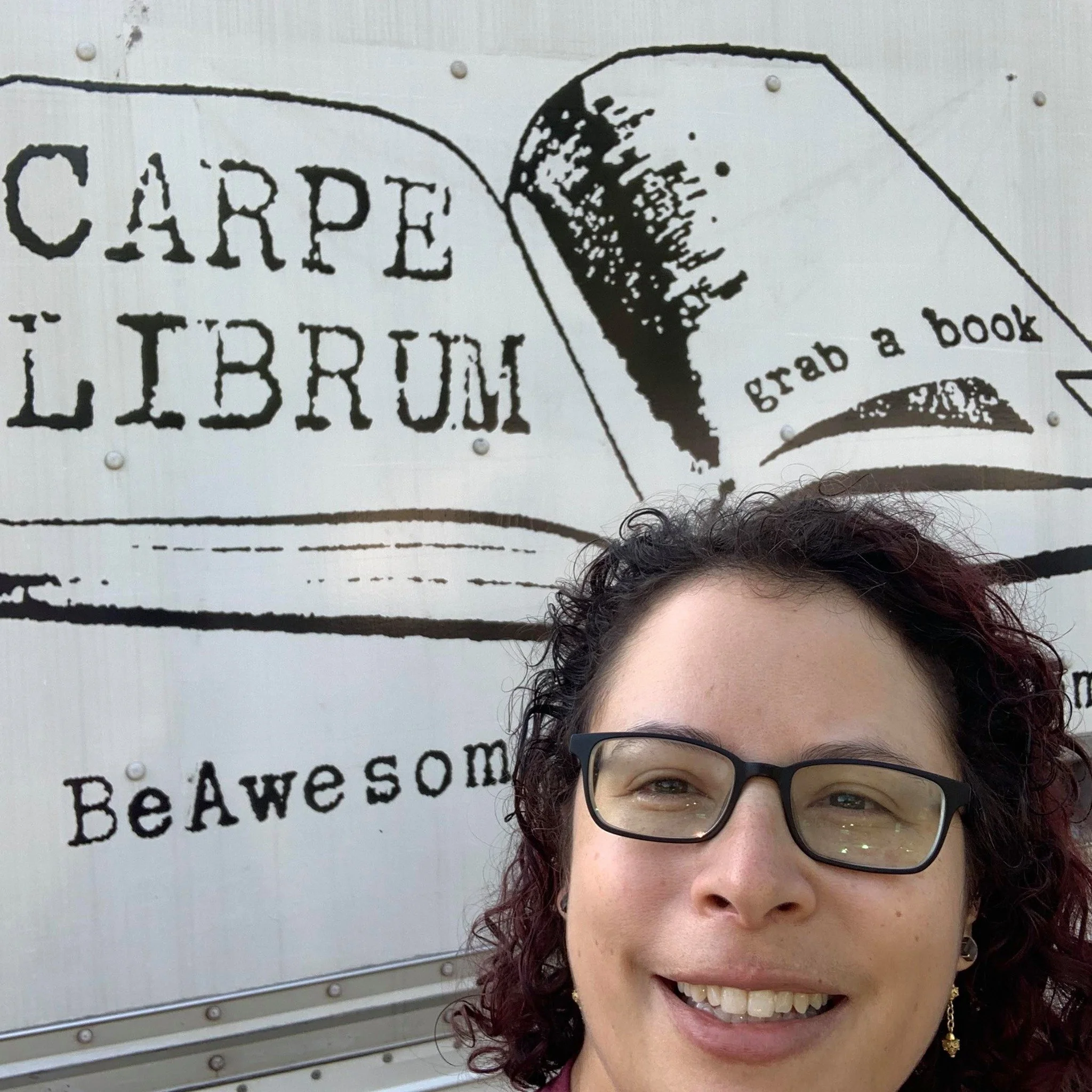 Romy Natalia smiling in front of a wall with a sign of an illustration of a book with the phrase 'grab a book.'