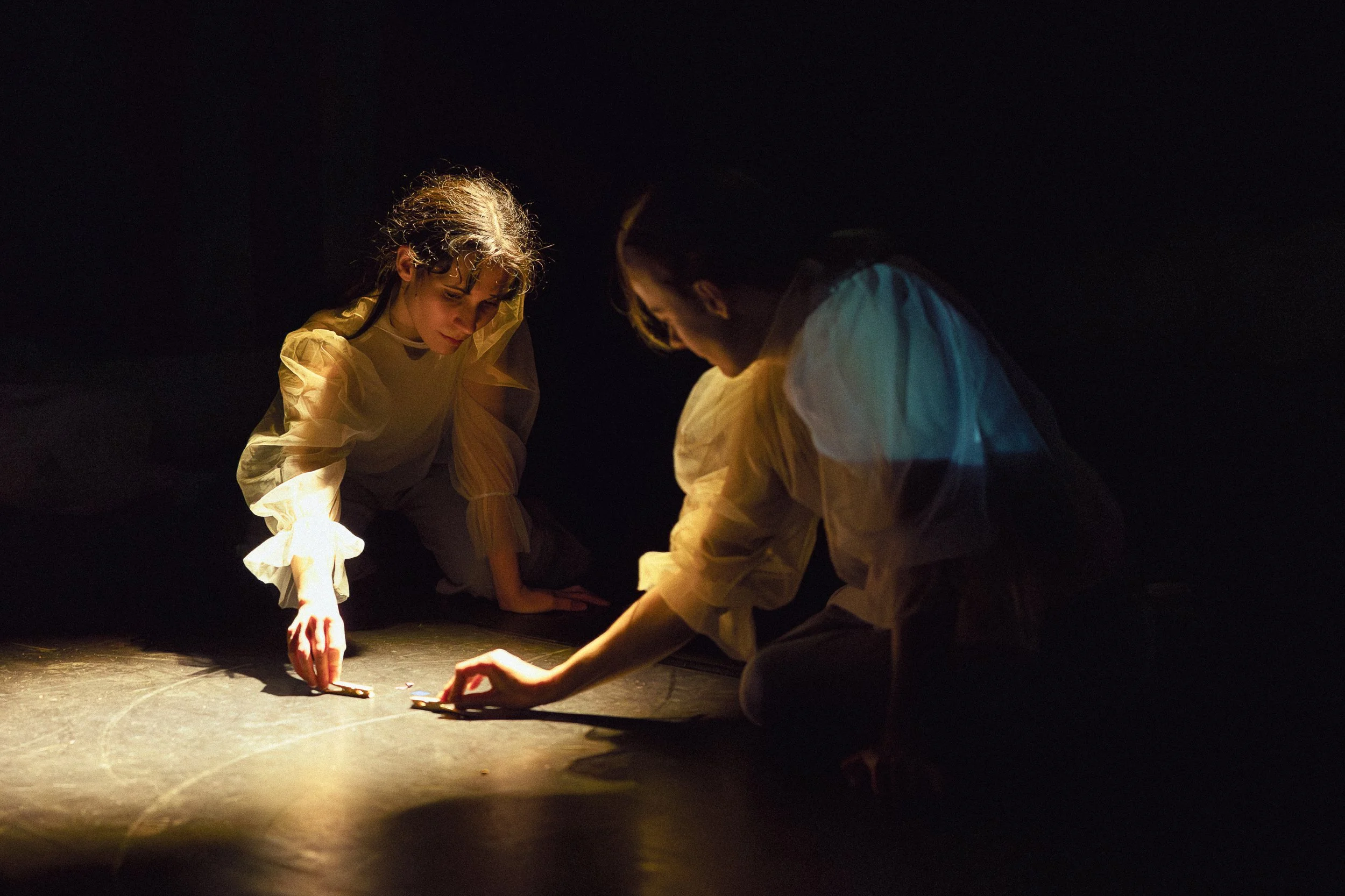 Two performers in yellow costumes kneel on a dark stage with a black background, illuminated by warm lighting, as they draw or write on the floor.