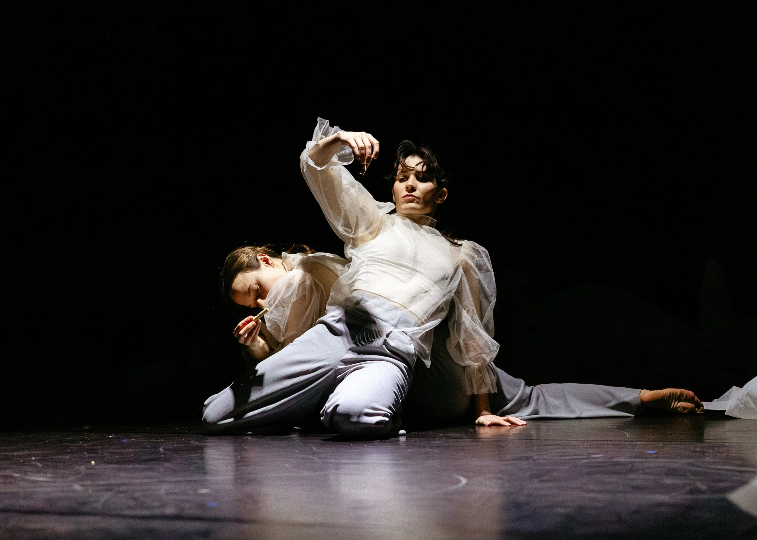 Two performers in white costumes practicing or performing a dance or theatrical piece on stage with a dark background.