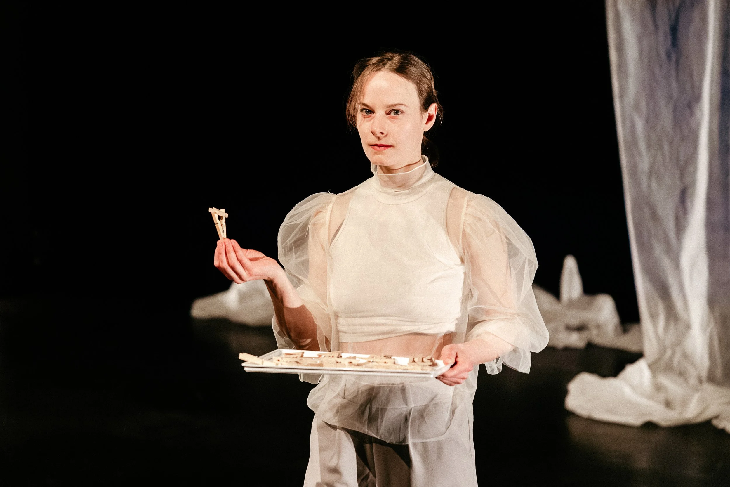 A woman in a light-colored, flowing dress holding a tray with clothespins on it, standing against a dark background with white draped fabric.