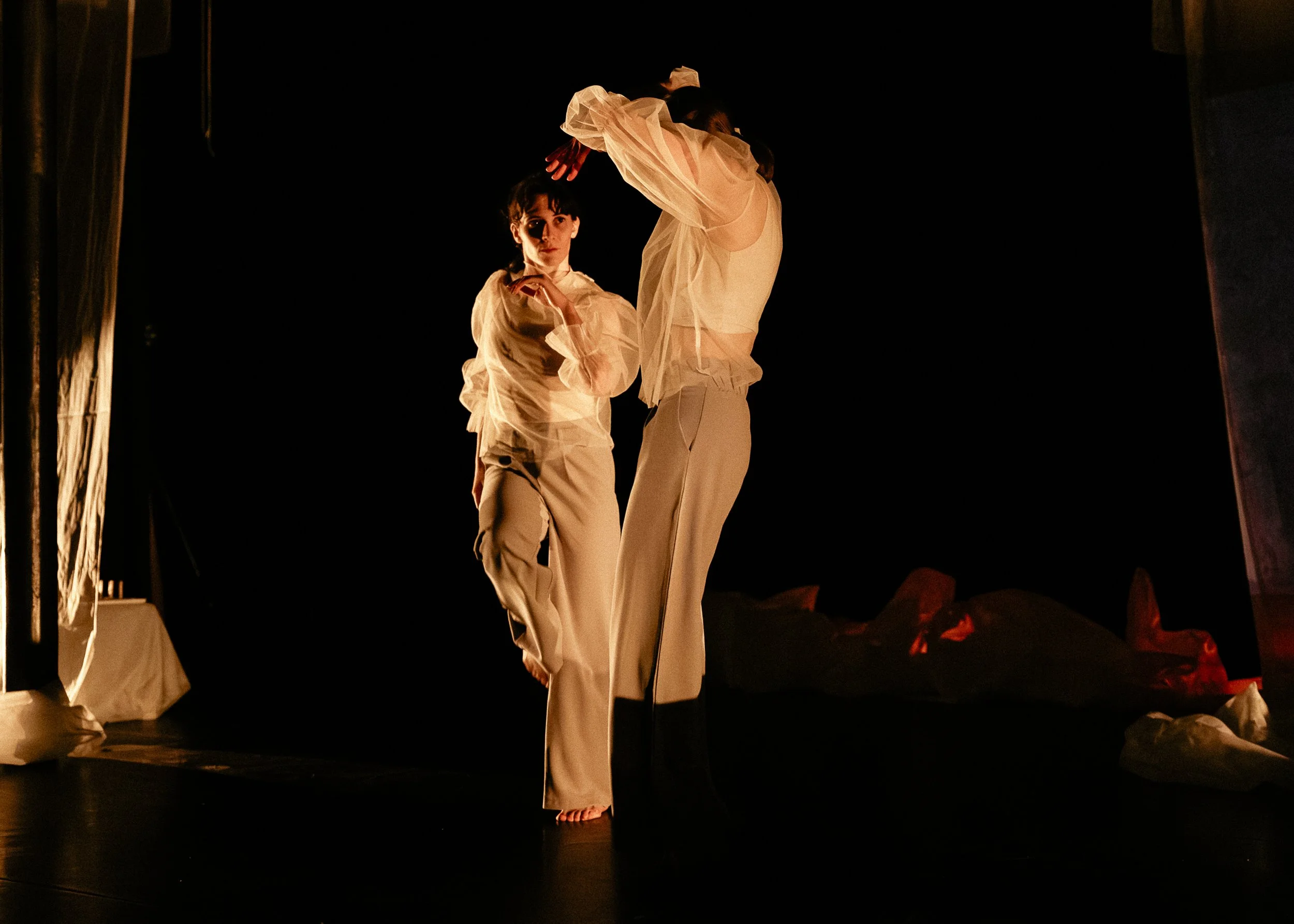 Two dancers perform on stage; one is a woman in a flowing dress, balancing on her toe with one leg raised high, and the other is a woman with short dark hair, standing with her hand near her chin, both illuminated by stage lighting against a dark bac