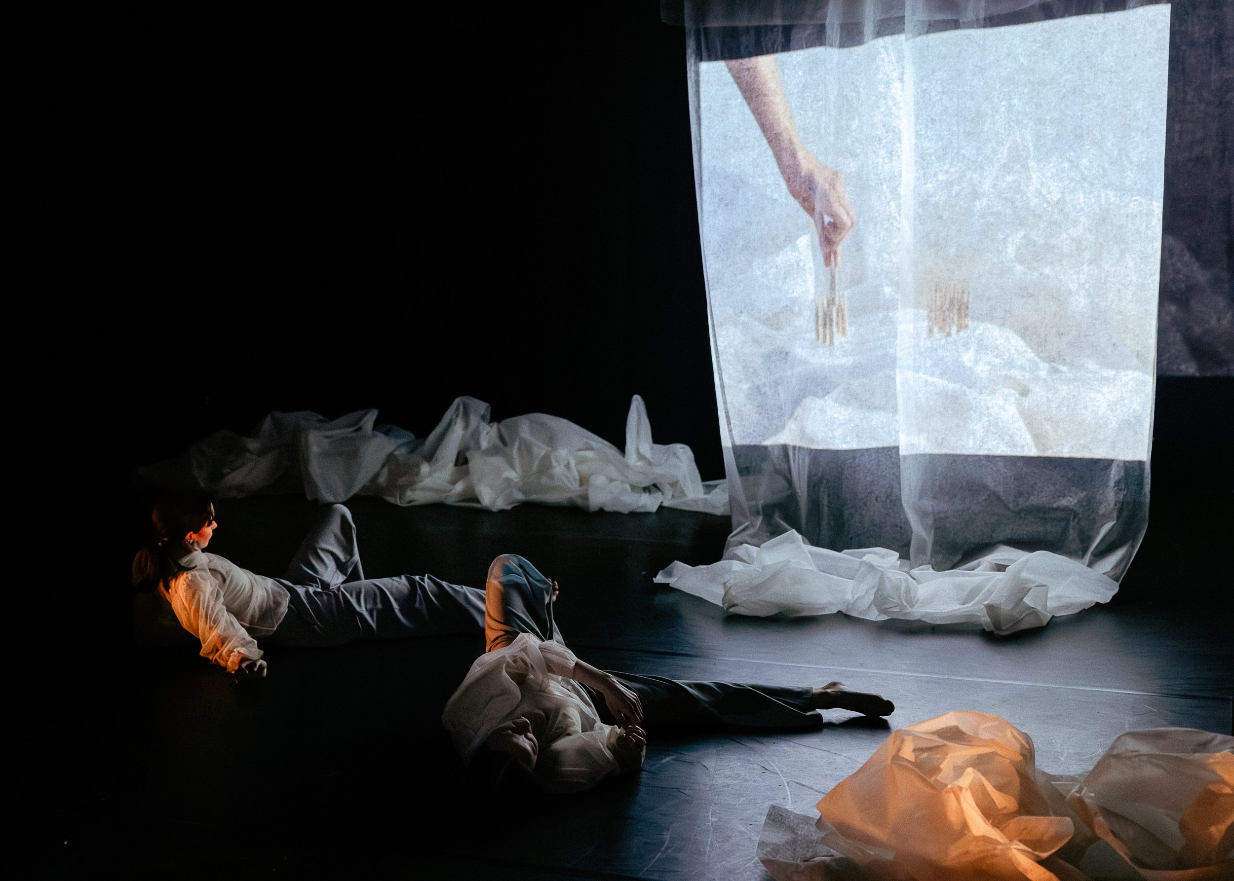 People in white costumes lying on the floor of a dark stage with scattered crumpled papers and a large transparent sheet displaying a hand holding brushes.