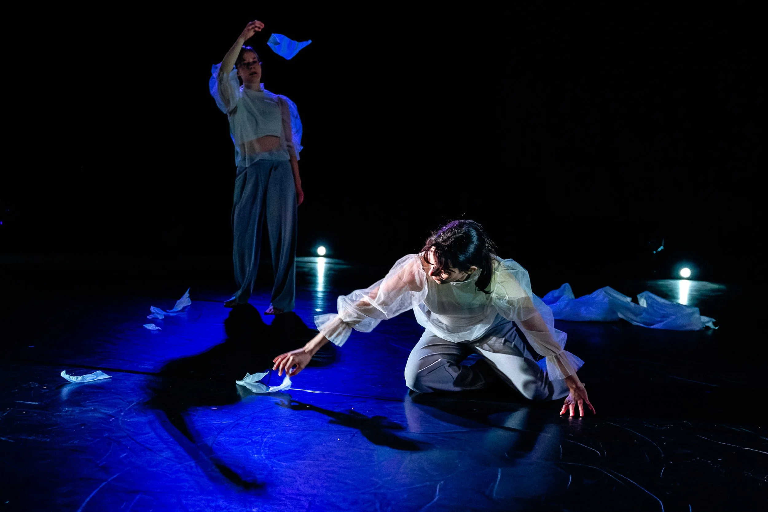 Two performers on stage, one kneeling and reaching out, the other standing and holding paper, with scattered papers on the floor, under dim blue lighting.