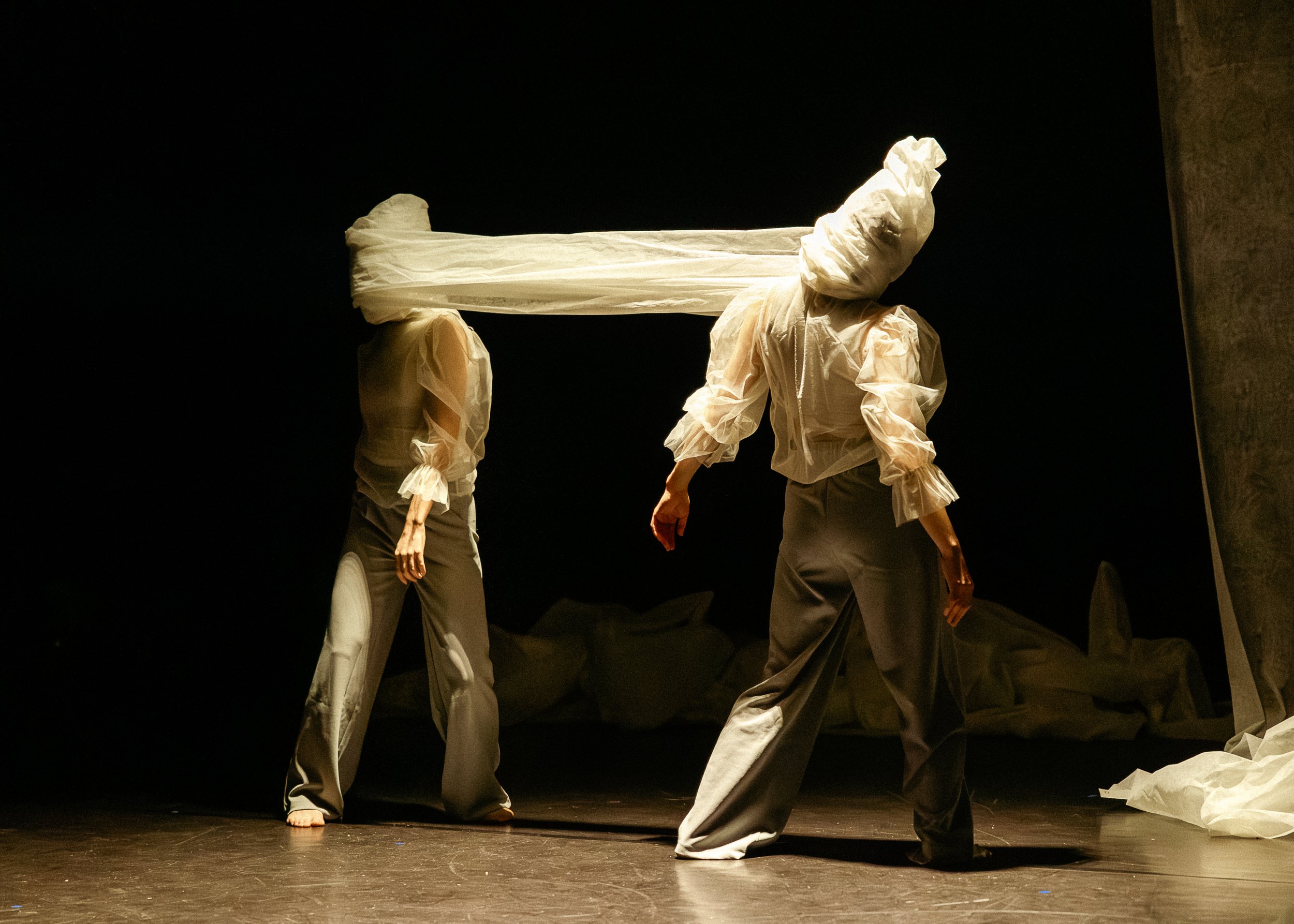 Three performers on stage wearing loose clothing and paper-like head coverings, with one performer kneeling and the others standing against a dark background.