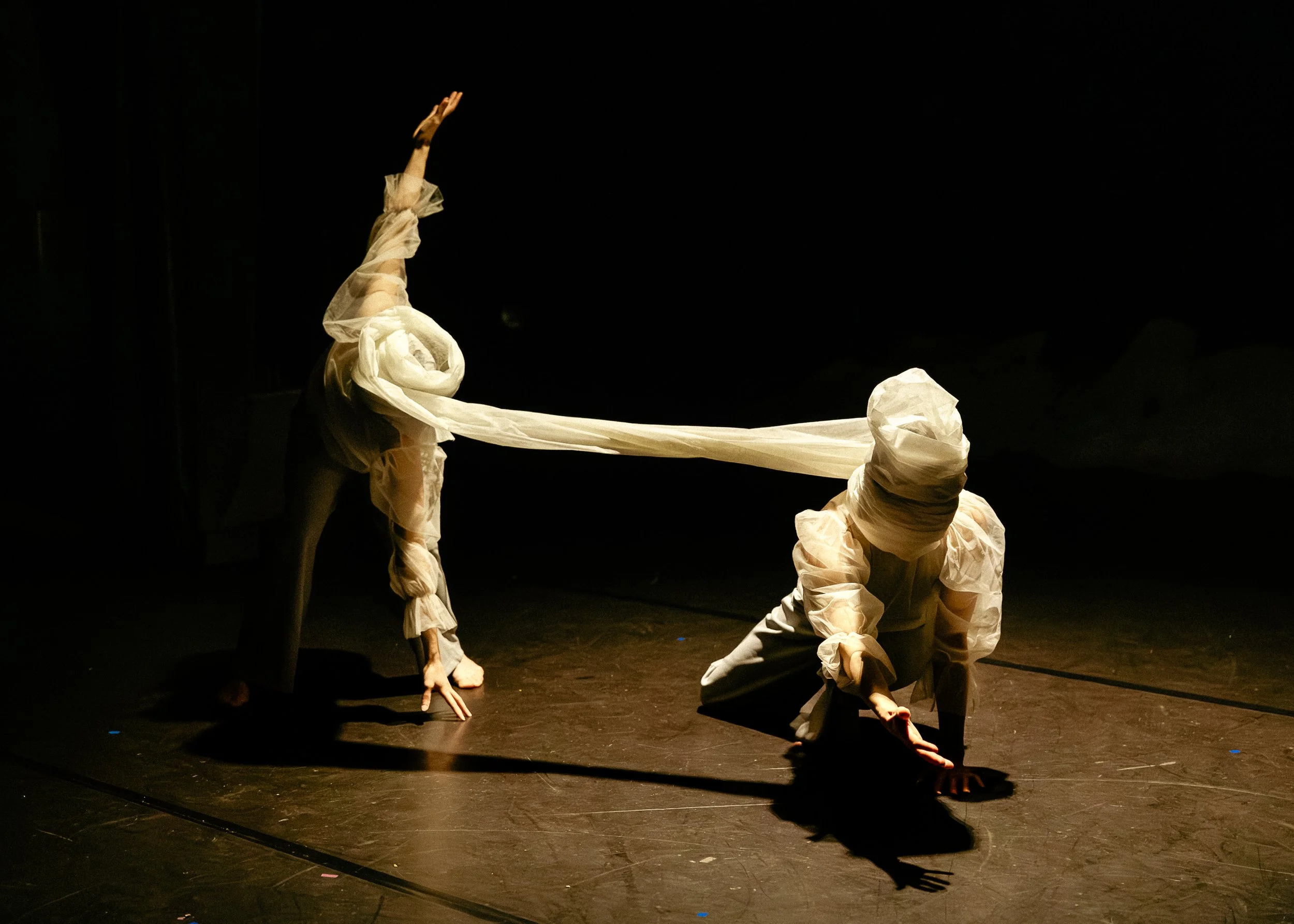 Two performers dressed in white fabric costumes are on stage, one are kneeling and the other is bent over, in a dark setting with minimal lighting.