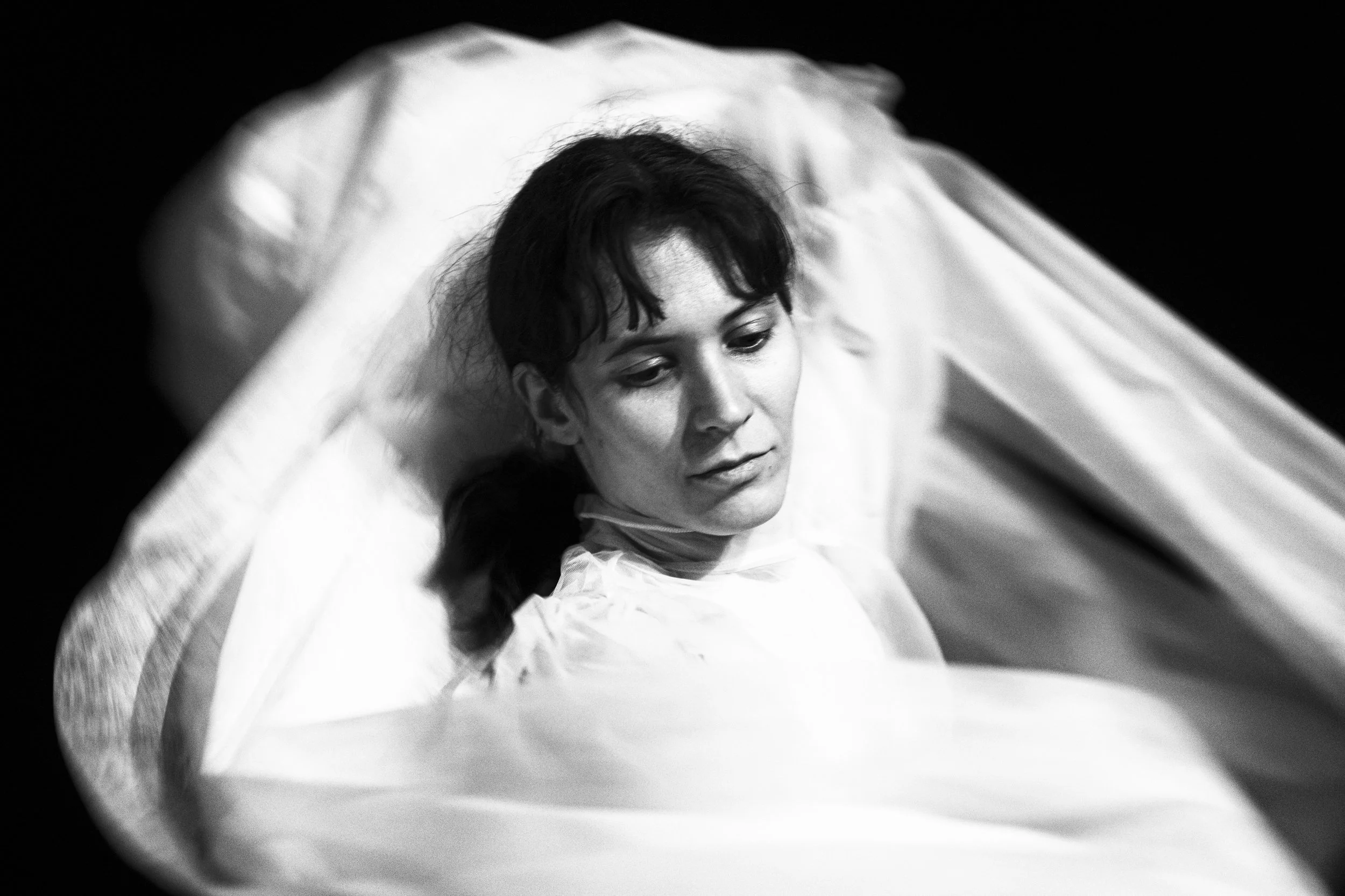 Black and white photo of a woman lying on a bed with her head resting on a pillow, looking downward with a contemplative expression.