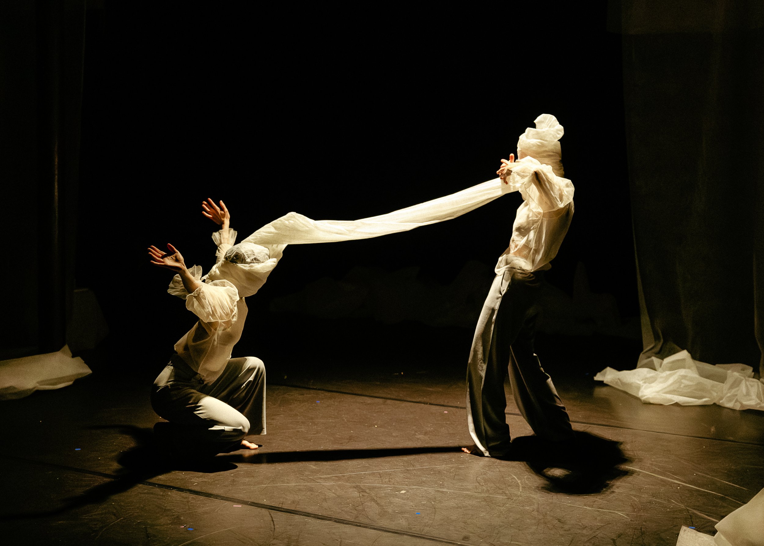 The image depicts four performers in white costumes with fabric wrapped around their heads and faces, engaged in a theatrical performance on a dark stage. They are positioned with one kneeling, one crouching, and two standing, with arms outstretched 