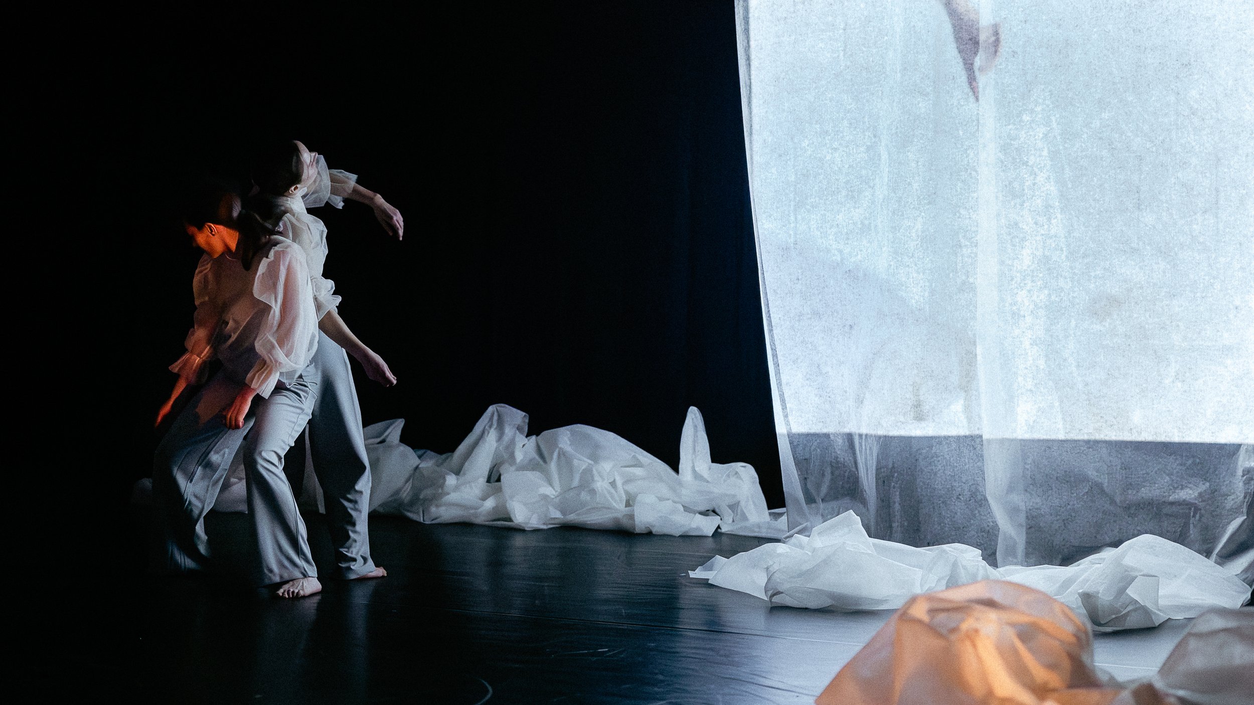 Performers in white clothing on stage, with crumpled paper and a large white sheet in the background, creating a minimalist artistic scene.