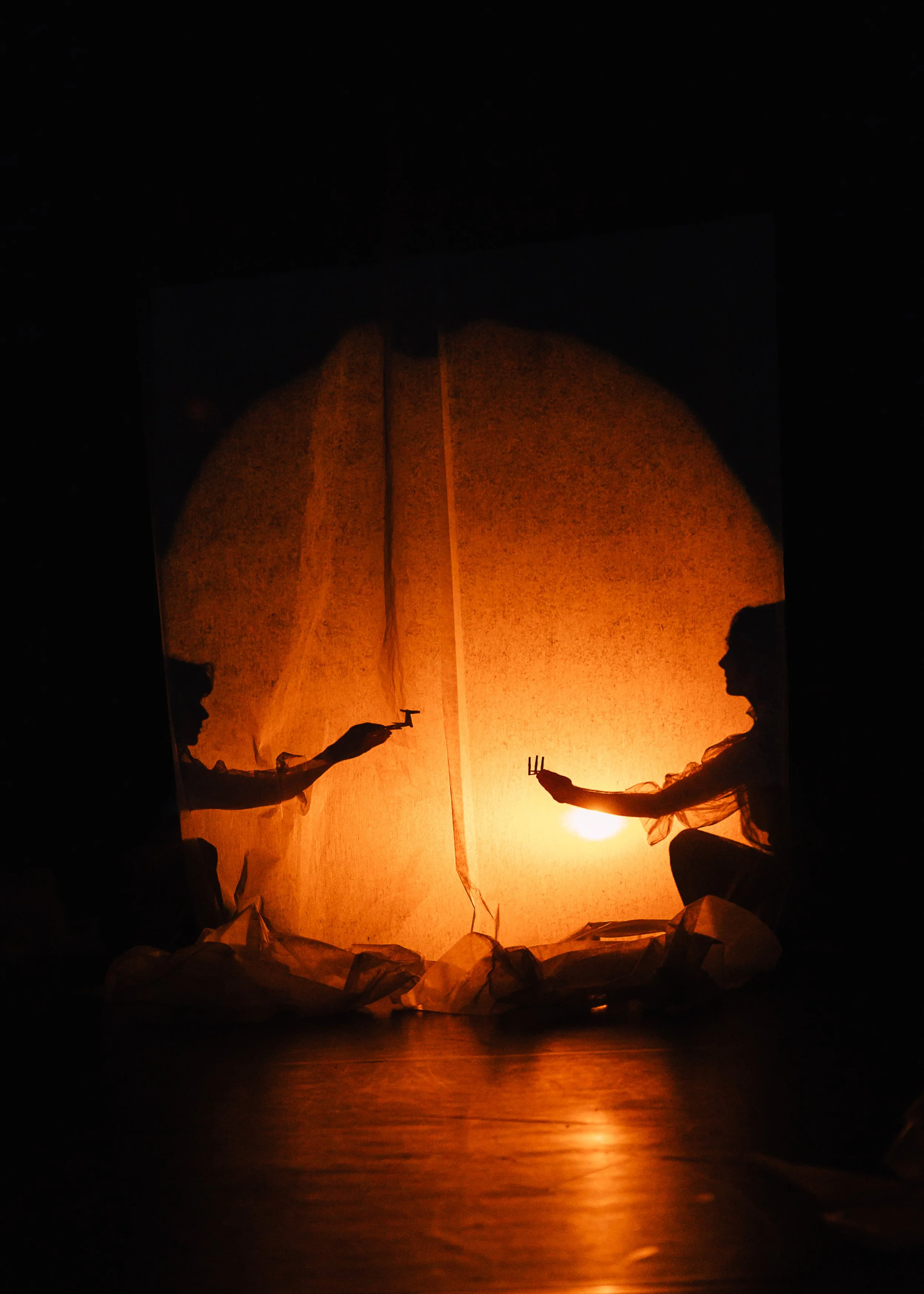 Silhouettes of two children blowing paper lanterns outdoors at sunset or sunrise, with a bright orange sky and the sun partially visible near the horizon. Crumpled paper surrounding them on the ground.