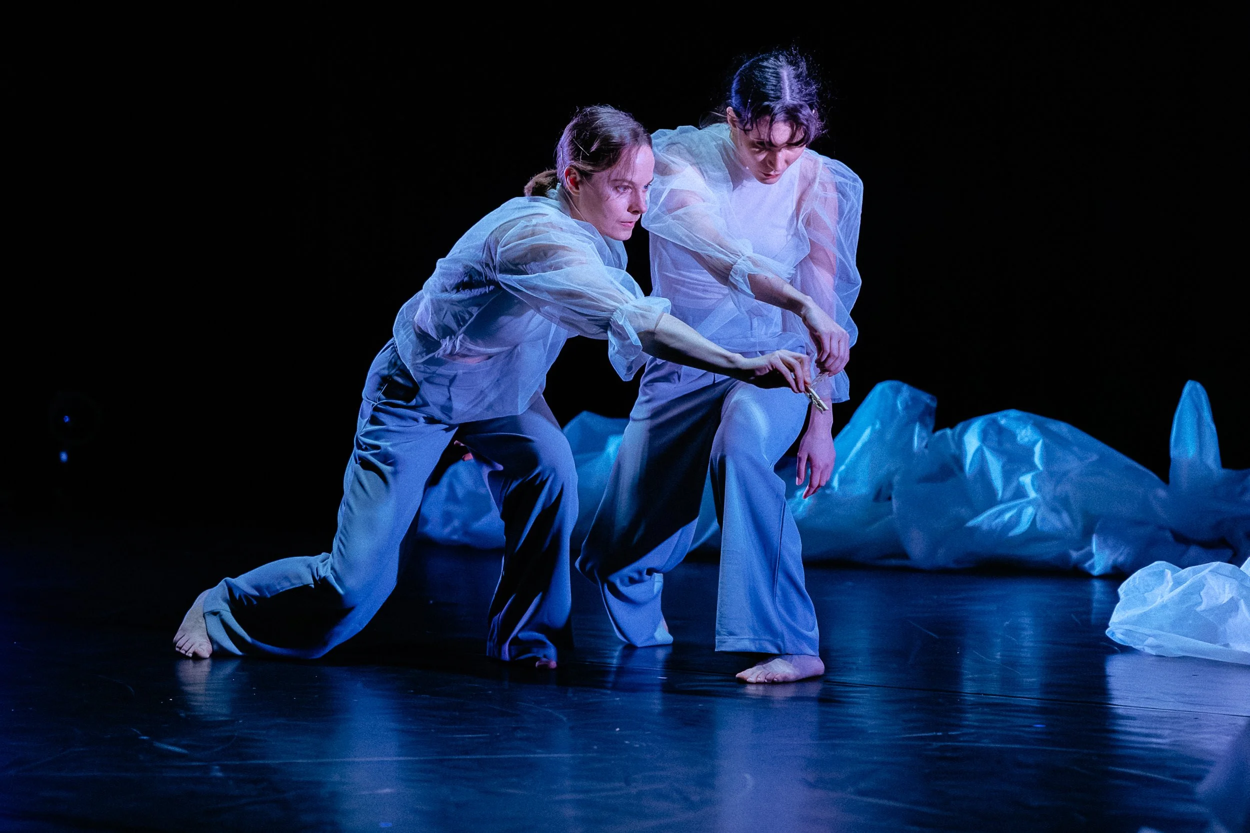 Two dancers perform on a dark stage, kneeling and leaning towards each other, dressed in translucent, white, puffy outfits, with crumpled white fabric on the floor.