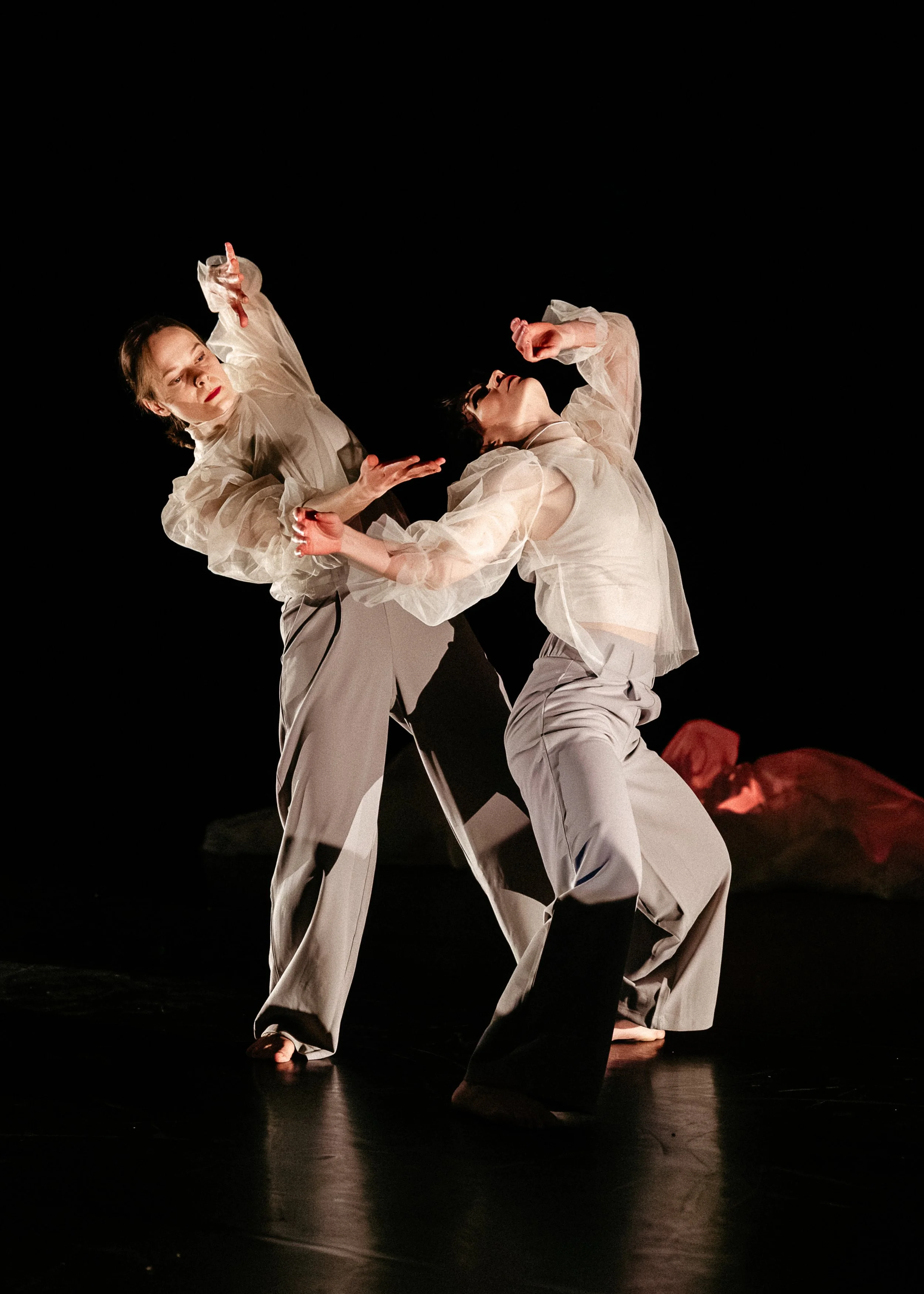 Two dancers in beige clothing perform on a dark stage, with one dancer standing and the other leaning back, holding hands.