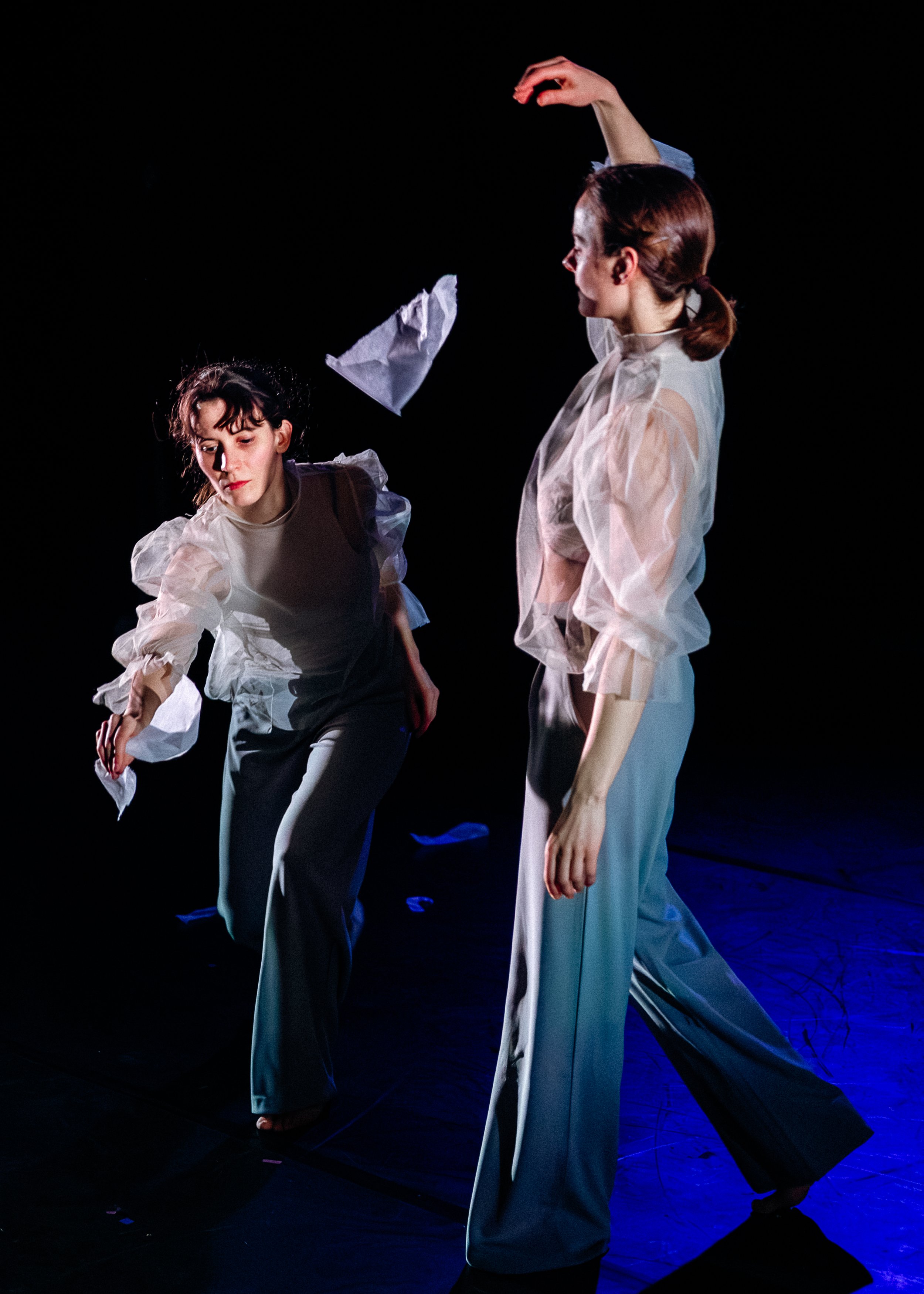 Two women perform a dance or theatrical piece on stage, illuminated by blue lighting against a dark background. Both are dressed in white blouses with puffed sleeves and dark trousers, with focused expressions and graceful poses.