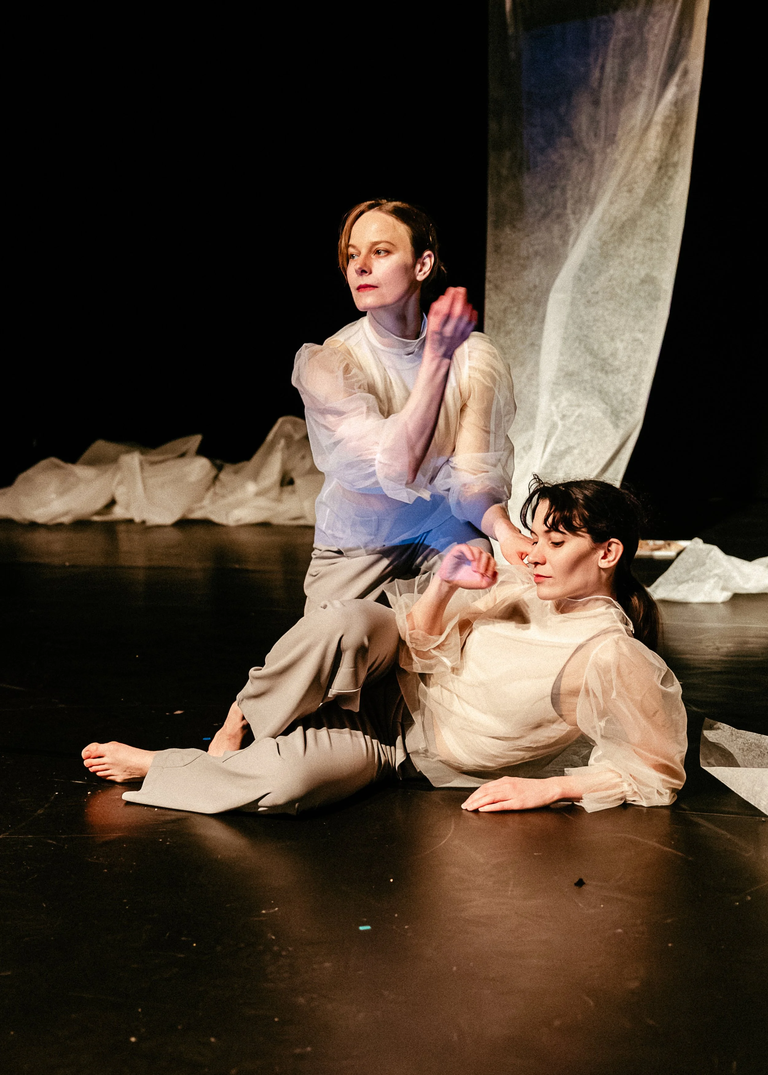 Two women with short dark hair, wearing beige and cream-colored costumes, are performing on stage. One woman is sitting on the floor with her eyes closed, while the other is kneeling beside her, touching her and looking away thoughtfully. The backgro