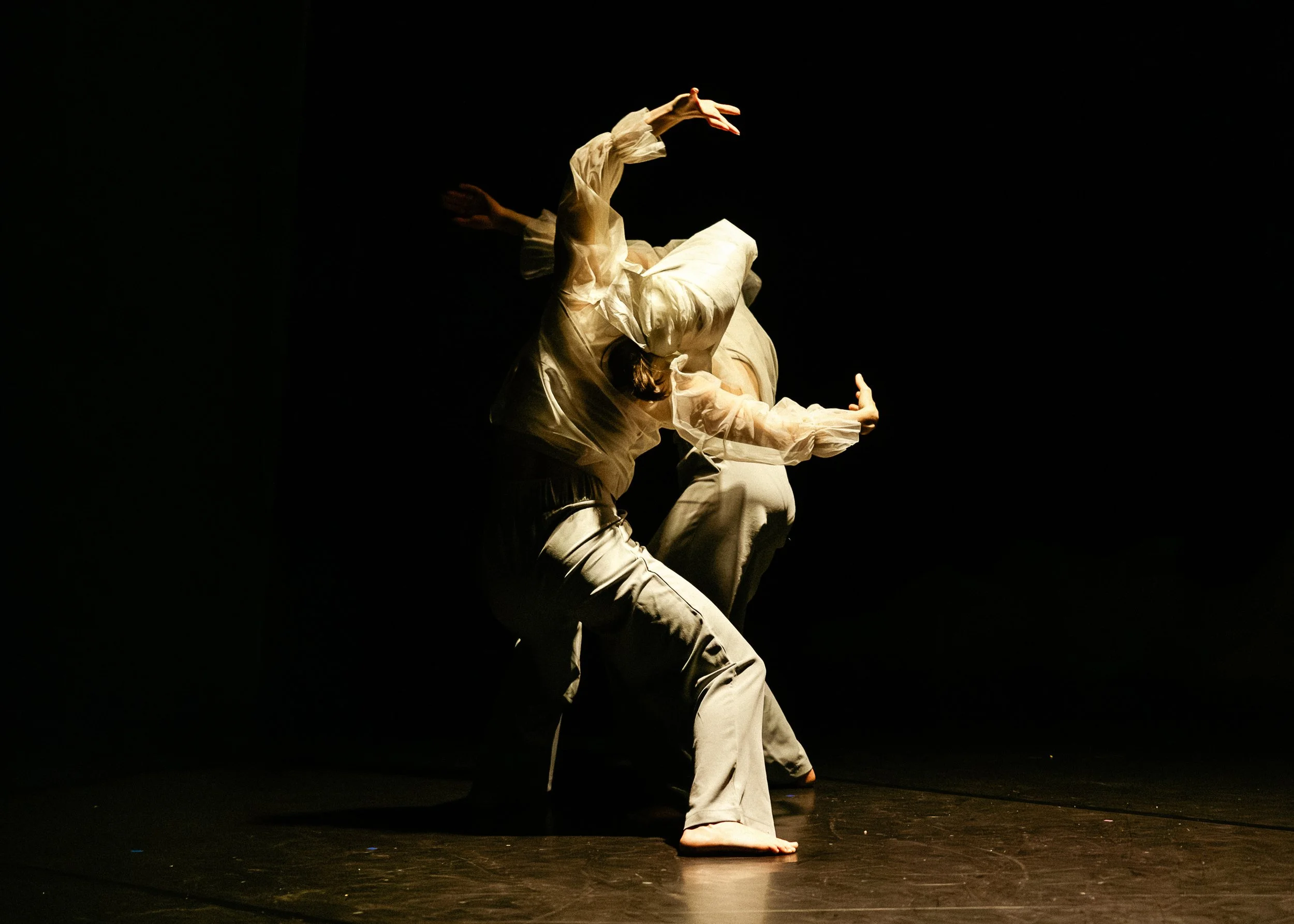 A performer dressed in flowing white garments dancing on a dark stage with dramatic lighting.