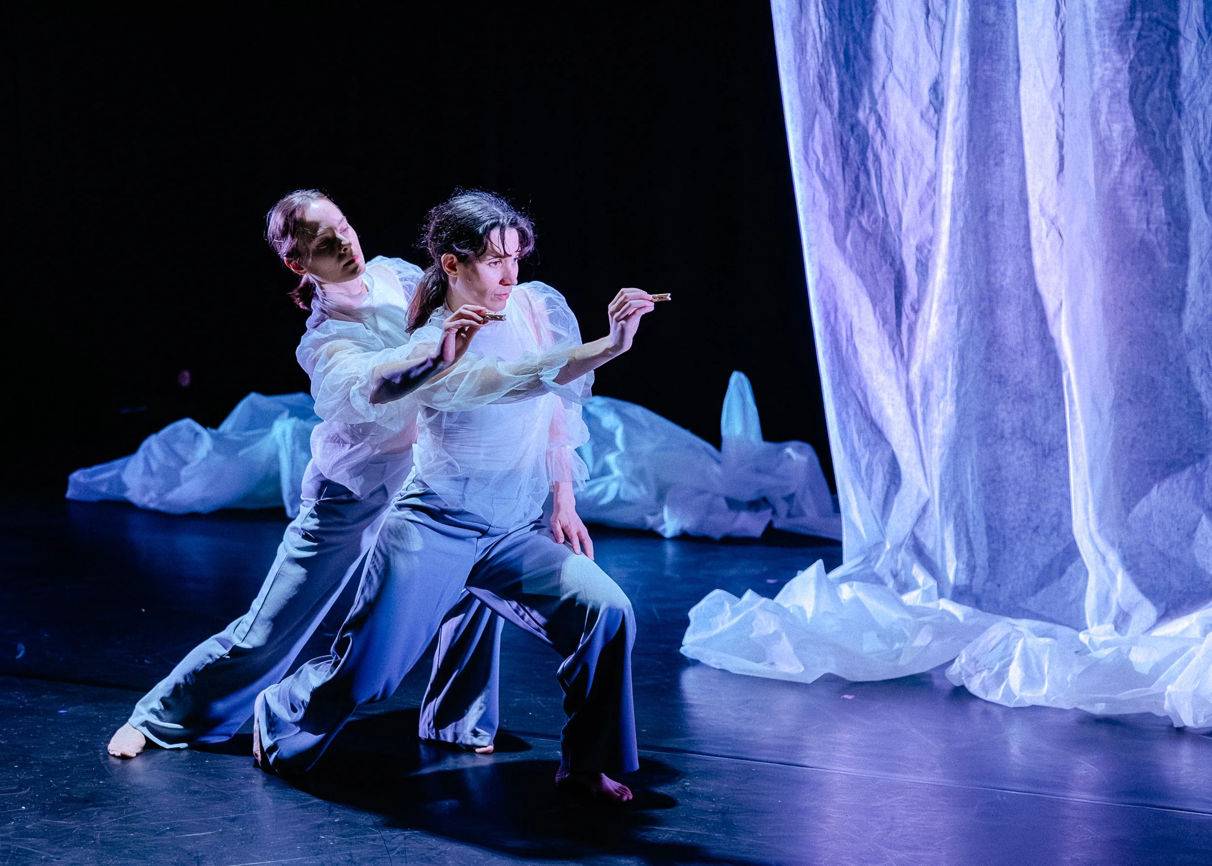 Two women performing a contemporary dance on stage with crumpled white and blue fabric and a dark background.