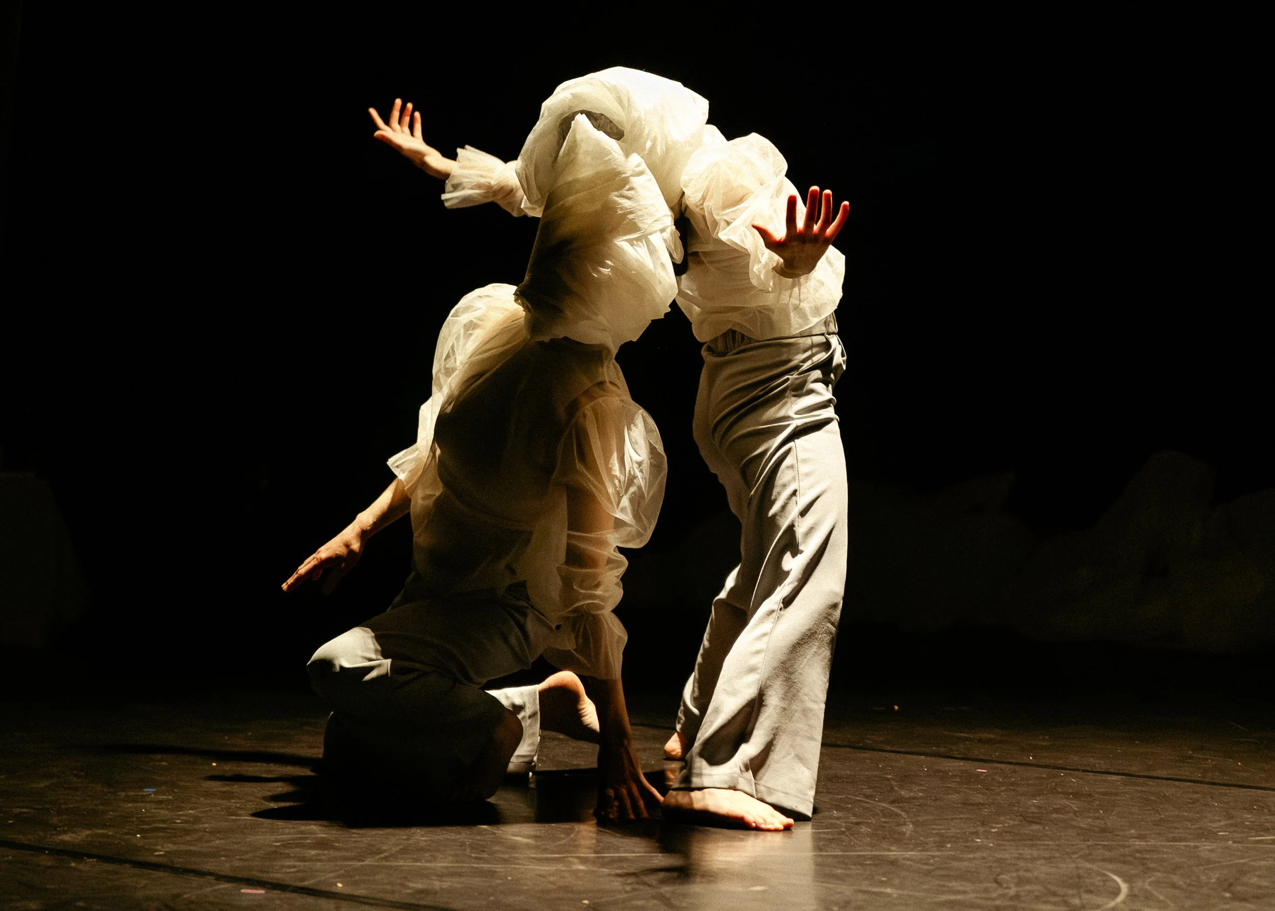 Performance of dancers on a dark stage, dressed in light-colored costumes, with one dancer kneeling and another standing, engaging in expressive movements.