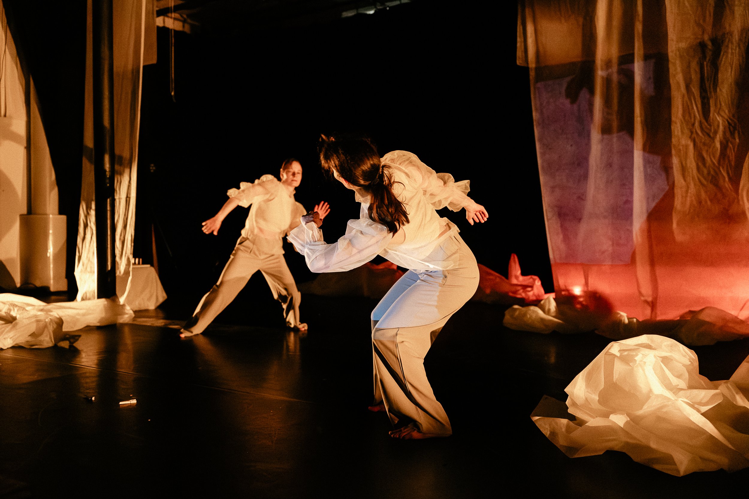 Two performers in white costumes are on stage, with one kneeling and the other standing, surrounded by crumpled paper props; stage is dimly lit with a dark background.