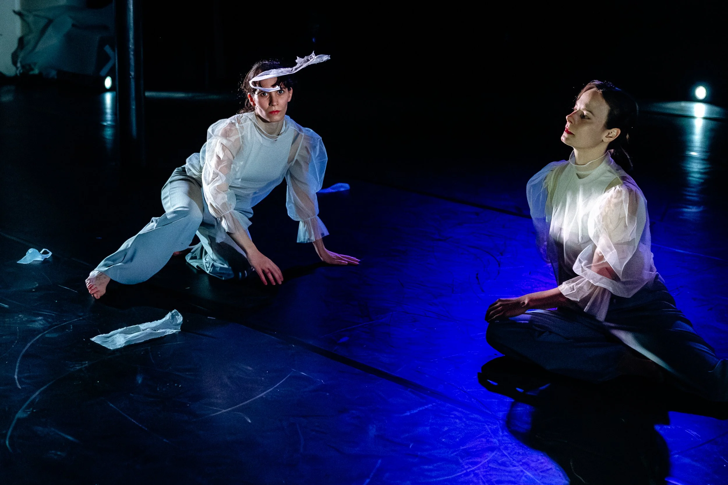 Two women performing on a dark stage, both dressed in white, kneeling with their hands on the ground, illuminated by blue stage lighting.