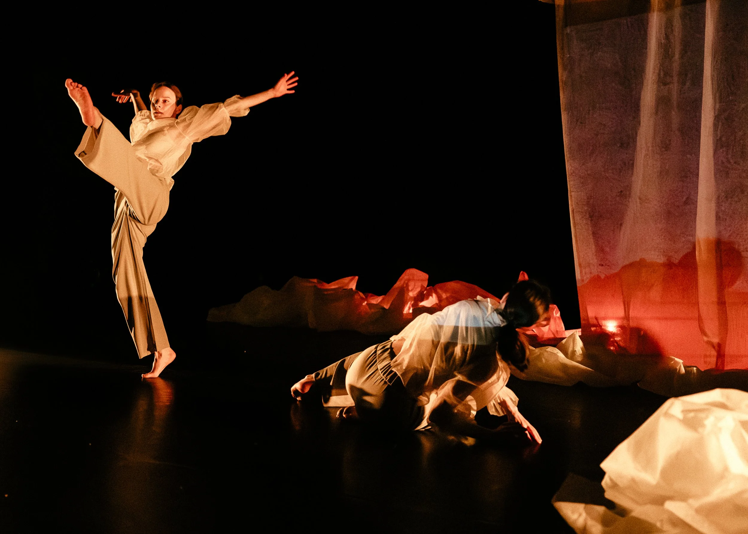 Performers dressed in beige costumes perform an expressive dance or theatrical piece on a dark stage, with some on the ground and one airborne with arms outstretched.