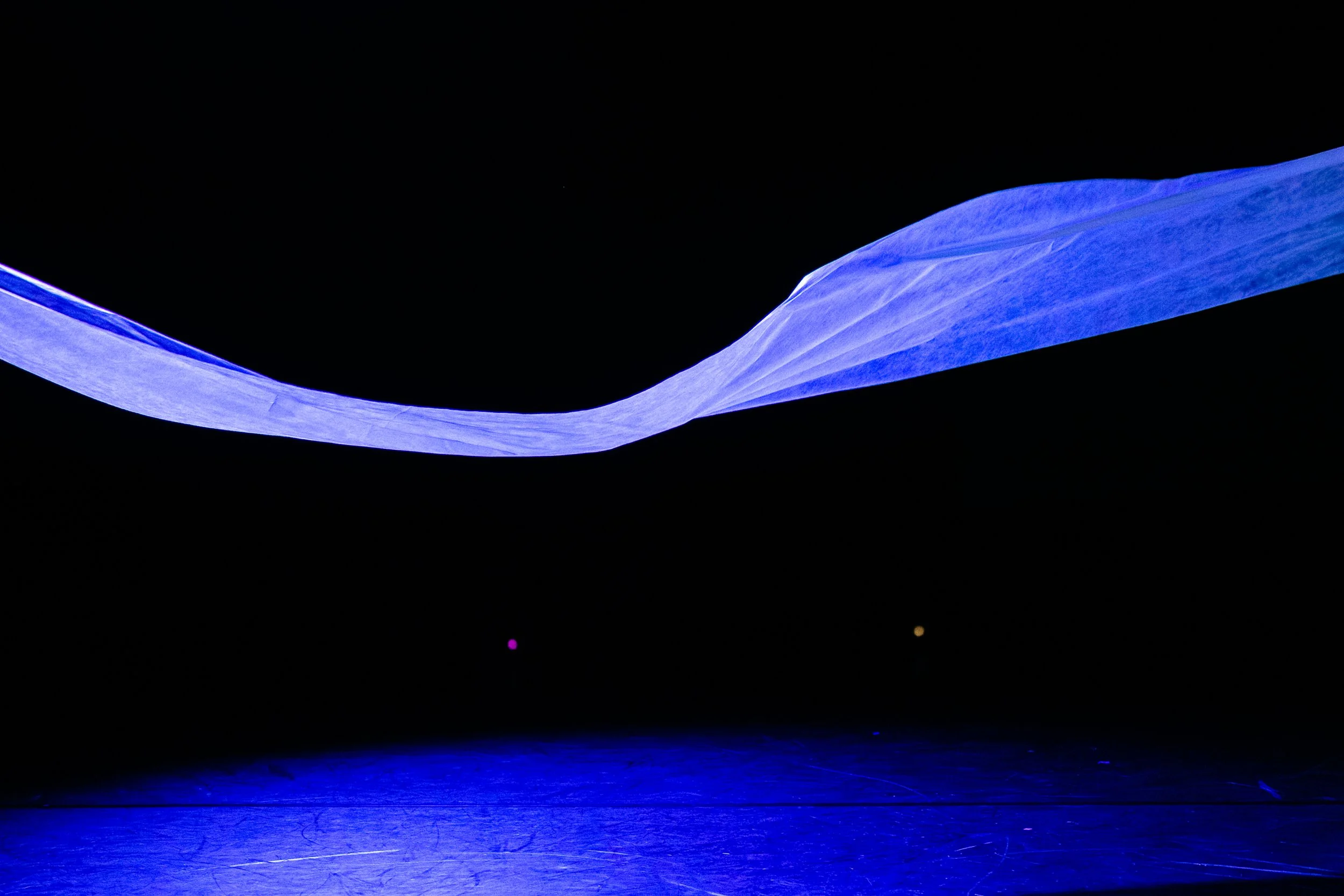 A dark stage illuminated with blue light, with a flowing, fabric-like installation suspended above.