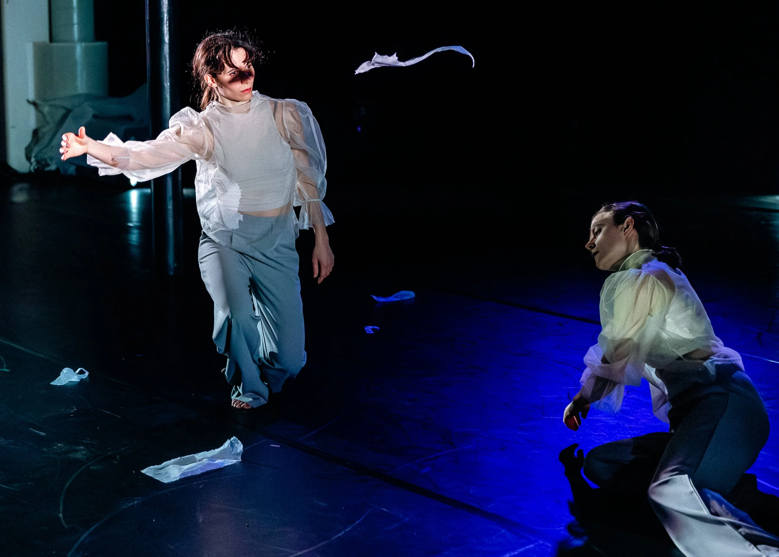 Two women in white costumes on a dark stage, one standing with arms outstretched and the other sitting on the floor, with scattered pieces of white paper or fabric around them.