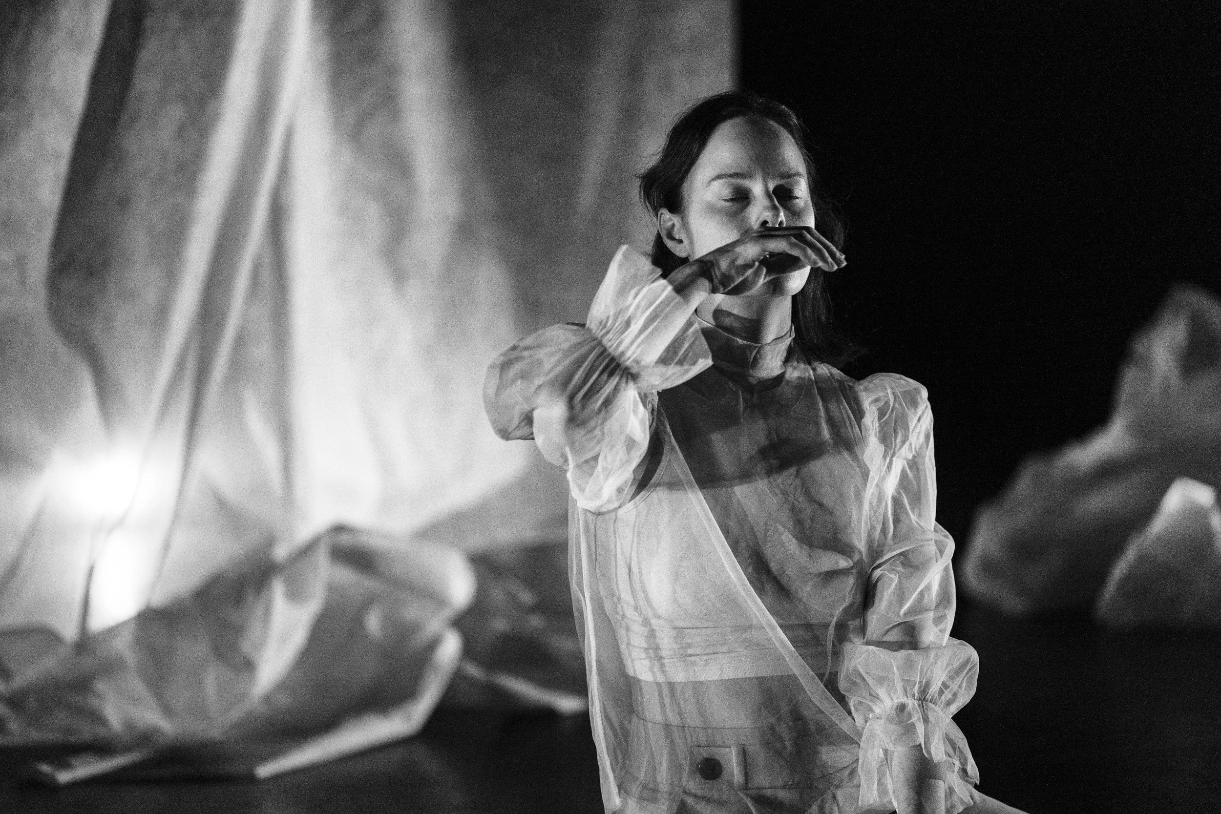 Black and white photo of a woman with closed eyes drinking from a glass, wearing a medical gown, with blurred figures in the foreground.