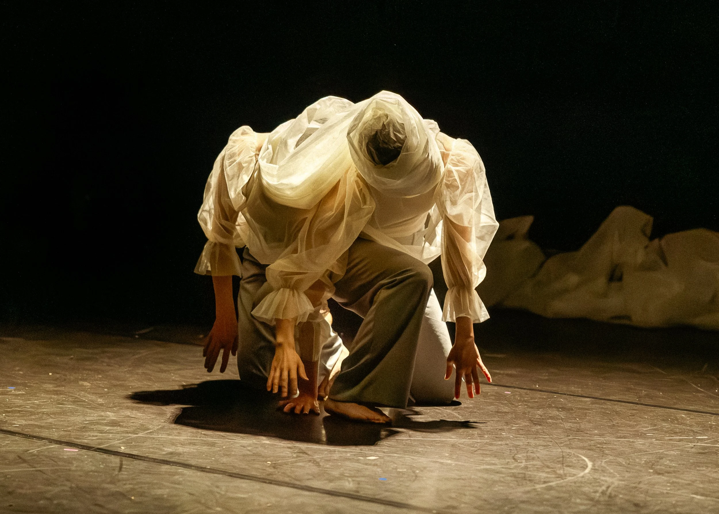 Performers in costumes are crouched on stage, posed with hands and fingers extended, facing downward, in a theatrical scene with a dark background.