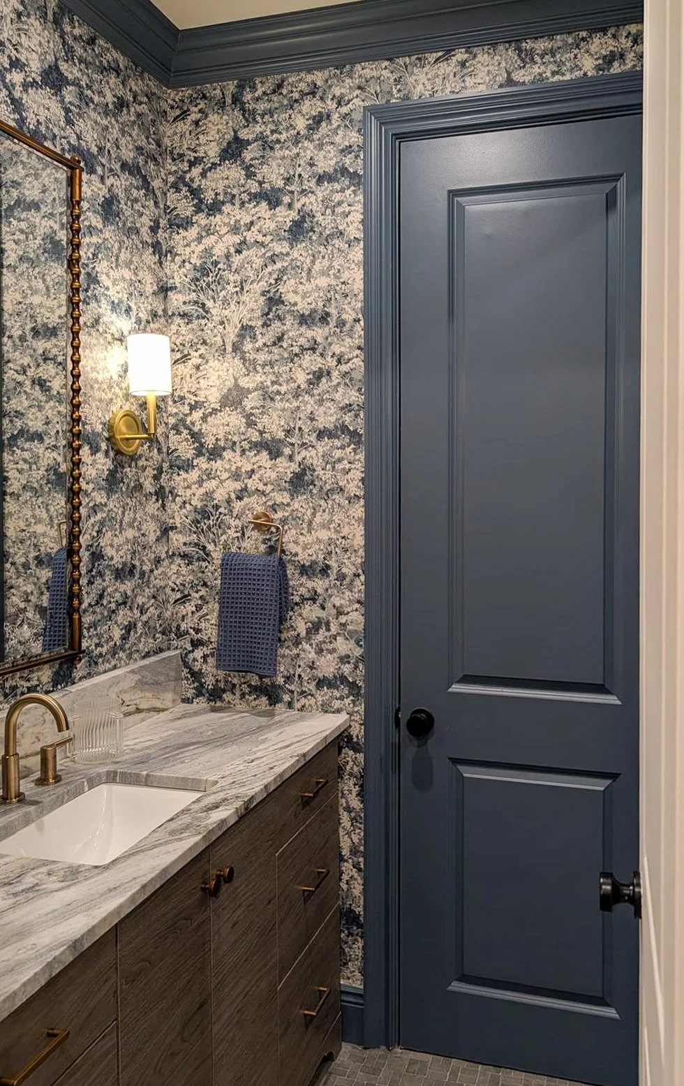 New Jersey jack and jill powder room with a vanity zone and separate toilet room, featuring indigo woodsy wallpaper, matching trim, brass accents, and a rustic stone mosaic tile floor.