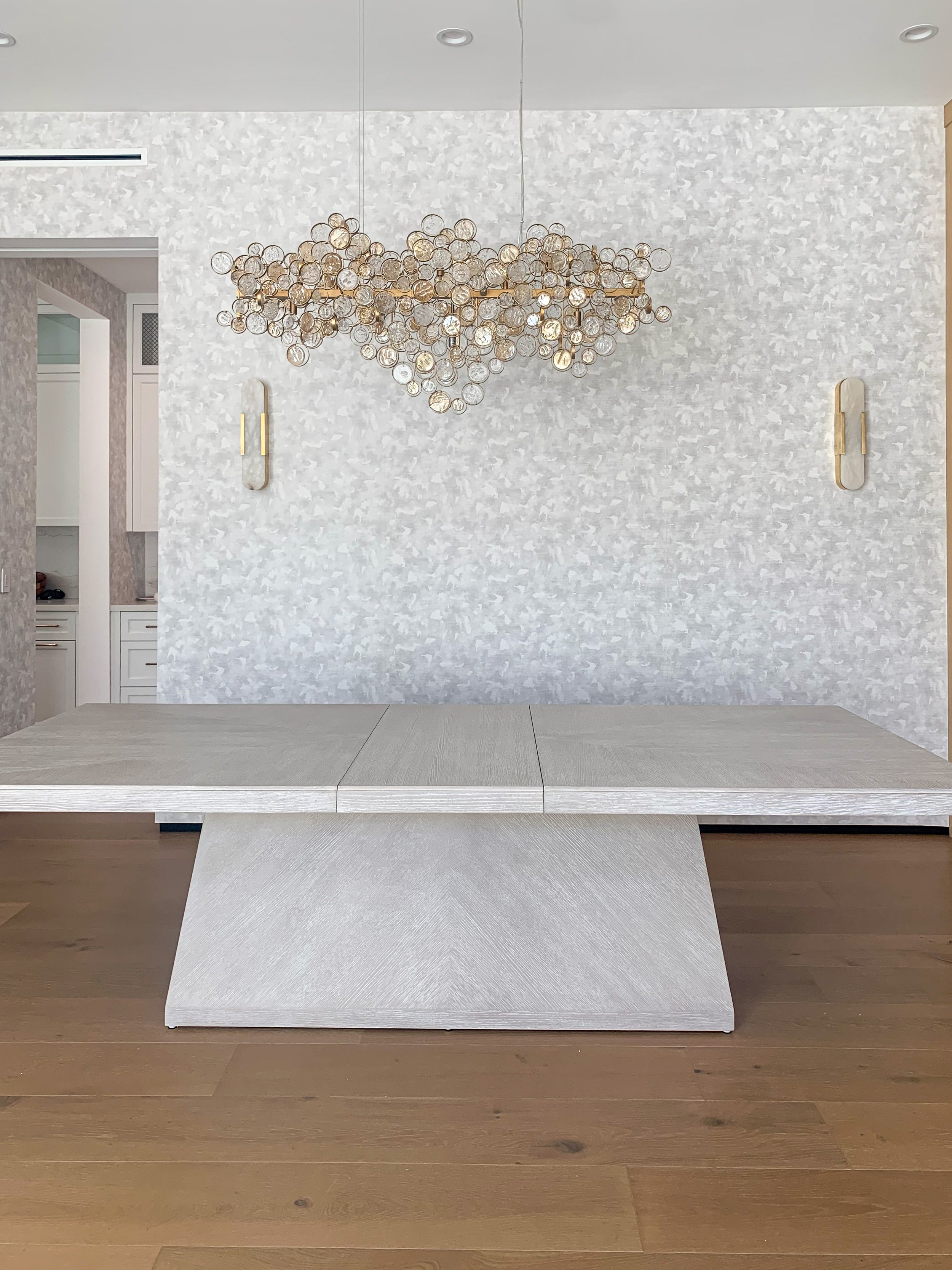 Dining room with cerused oak dining table, brass and glass linear chandelier, alabaster and brass pendants, and gray textured wallpaper.