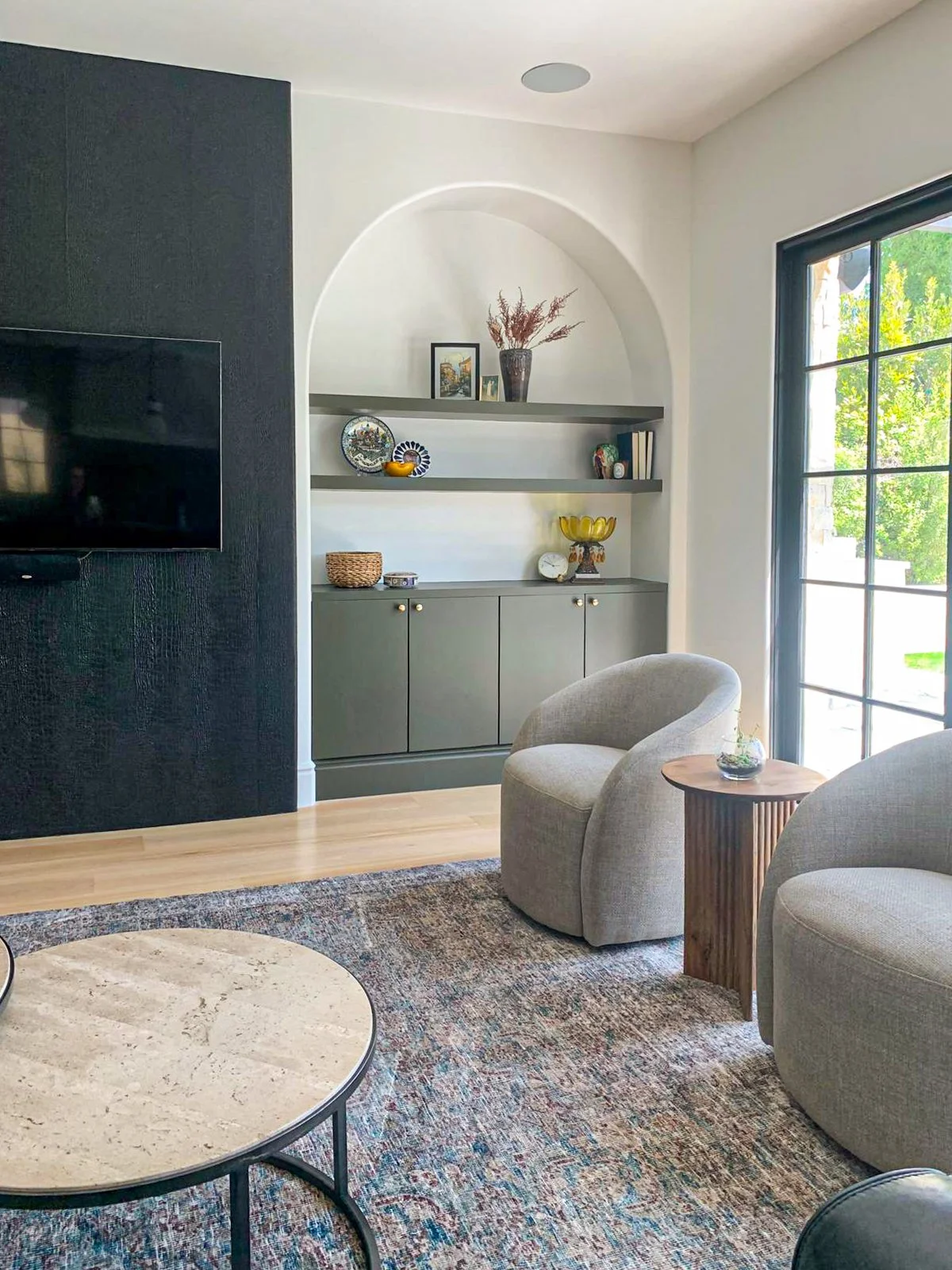 Beverlywood family room with arched niche shelving, green cabinetry, black plaster media wall, mounted TV, and a wall of pivot doors for indoor-outdoor living.