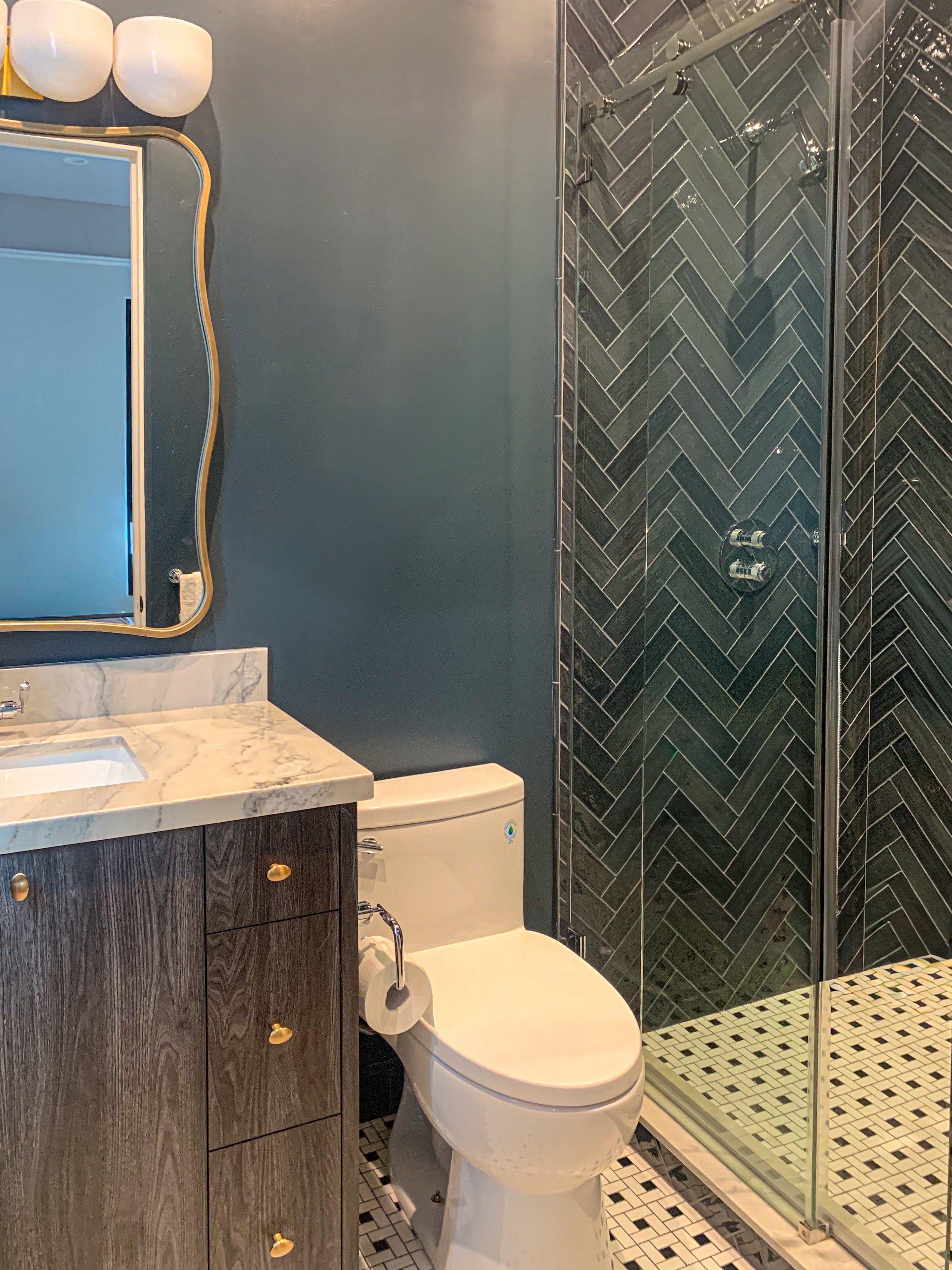 Navy color-drenched bathroom: herringbone subway tile, frameless shower enclosure, chrome/brass fixtures, vanity mirror and lighting, marble countertop and accents, and pinwheel mosaic floor.