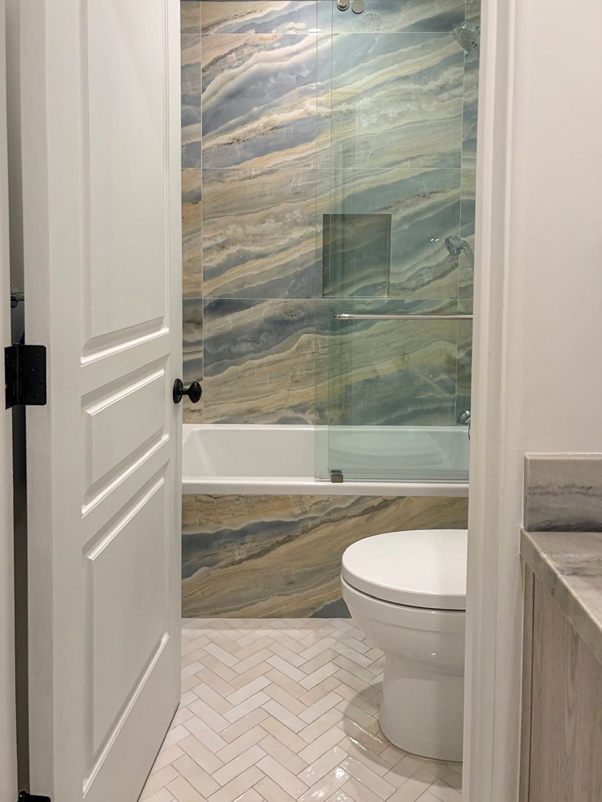 En suite guest bathroom in Beverlywood featuring a blue and beige faux marble tiled bathtub alcove with a frameless glass enclosure and a herringbone patterned tile floor.