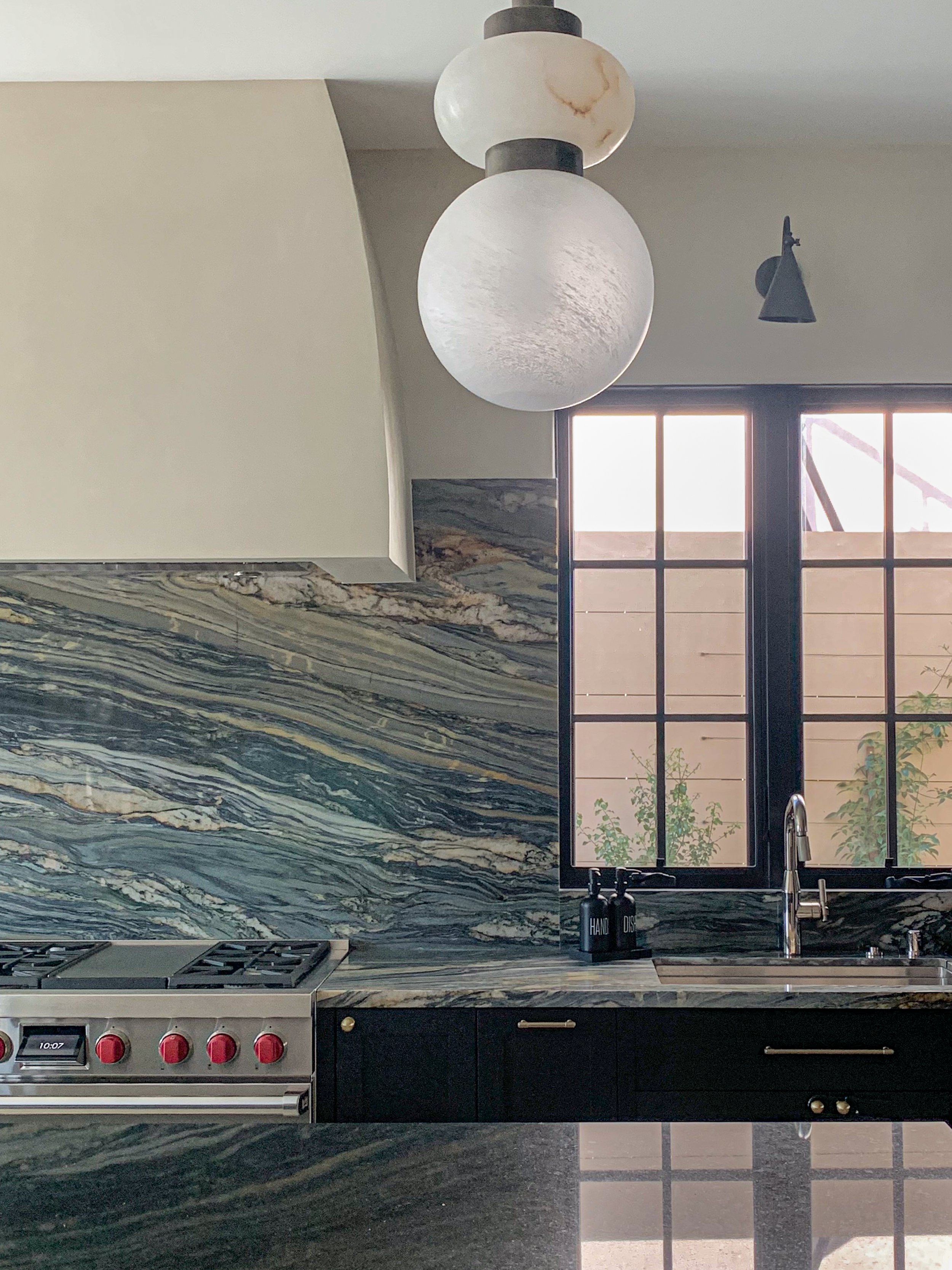 Moody black kitchen with quartzite countertop and backsplash, plaster hood, polished nickel faucets, brass cabinet hardware, stainless steel range, bronze sconces, and alabaster pendant lights.